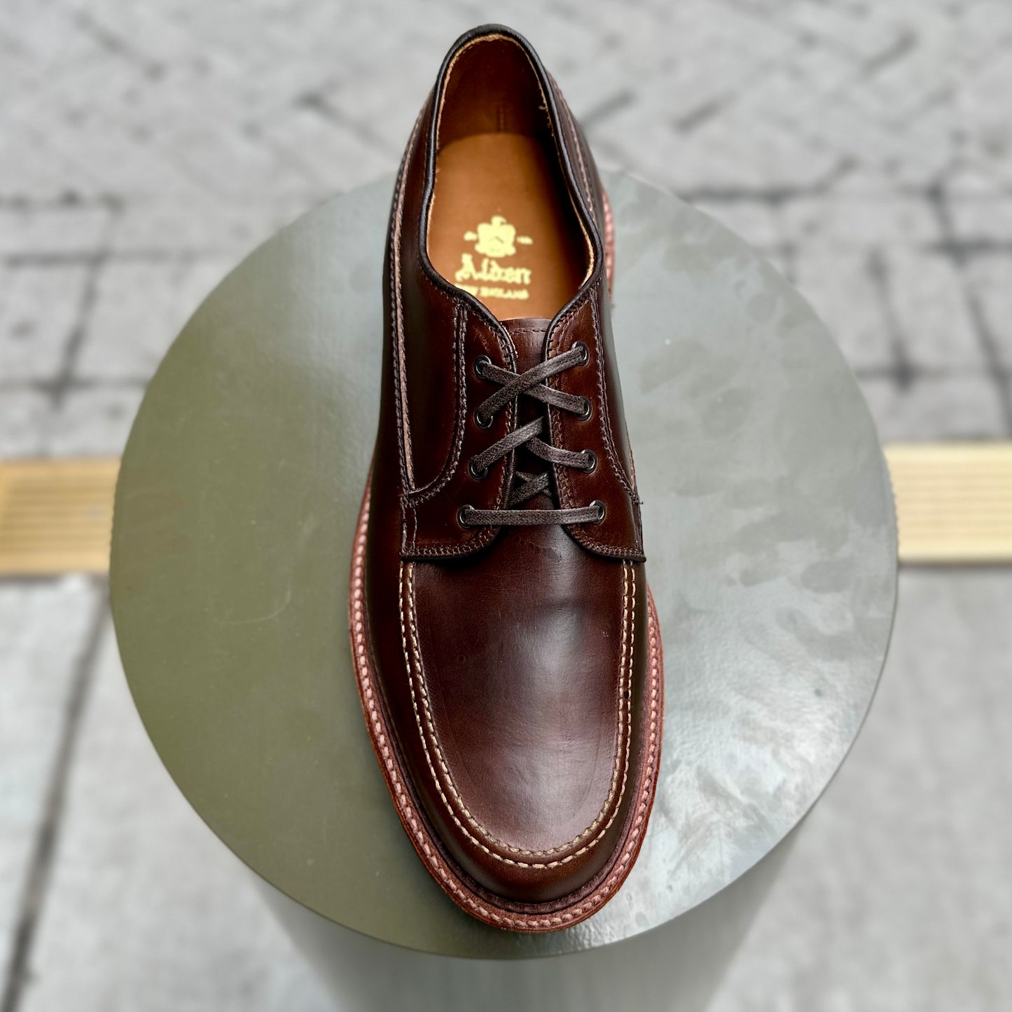 Displayed on a round pedestal, the D3616C - Ranger Mocc in Brown CXL by Alden Madison features a Commando Sole, detailed sole stitching, and a brand logo on the insole. The blurred tiled backdrop enhances its elegance.