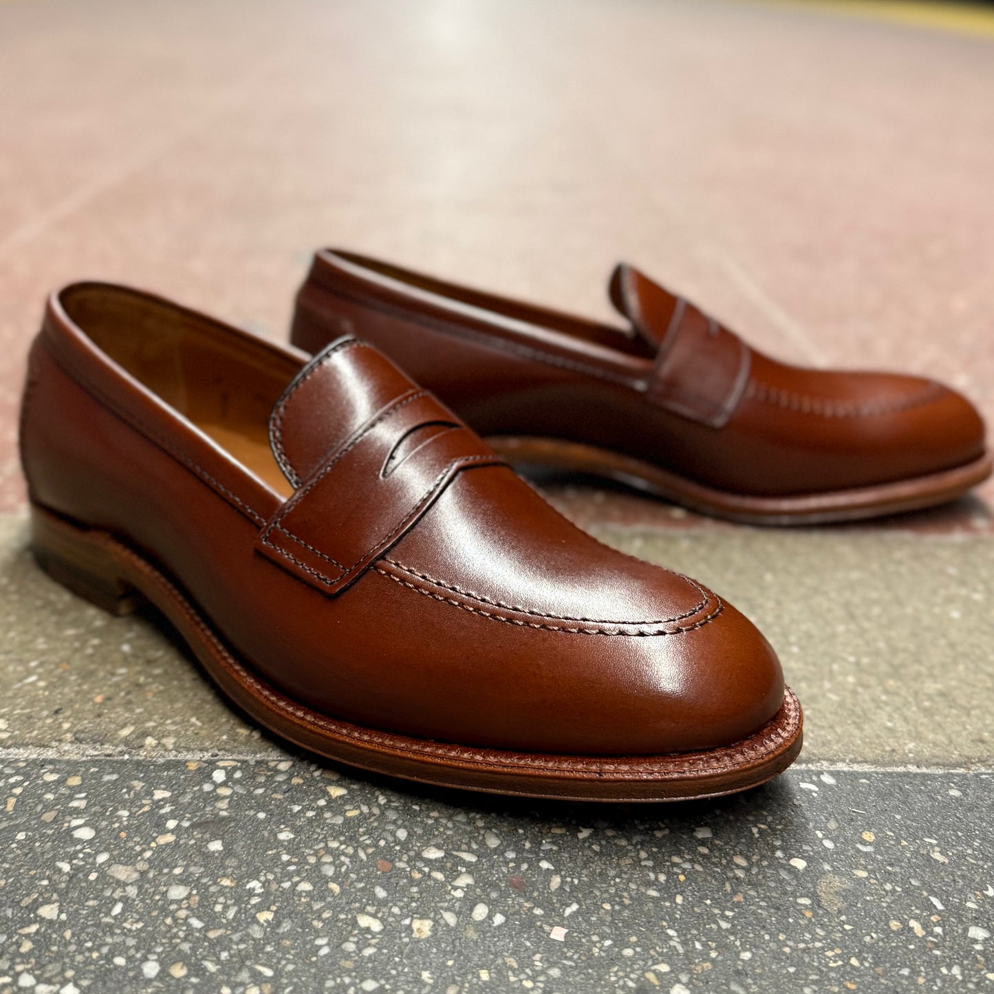 A pair of Alden Madisons D1235 - Madison Penny Loafers in polished Madison Brown Calfskin with a sleek finish and decorative stitching on a speckled tile surface, featuring subtle top detailing and an elegant, classic design.