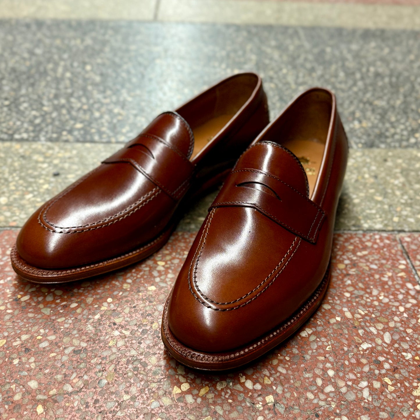 The sleek D1235 - Madison Penny in Madison Brown Calfskin by Alden Madison is gracefully positioned on a speckled, multicolored tile floor.
