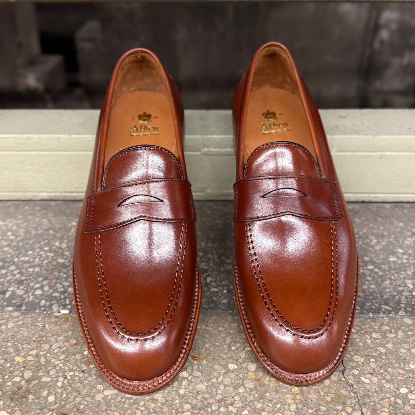 A pair of D1235 - Madison Penny loafers by Alden Madison in rich brown calfskin, featuring a leather sole, sleek stitching details, and a decorative strap, sit side by side on a stone-textured surface.