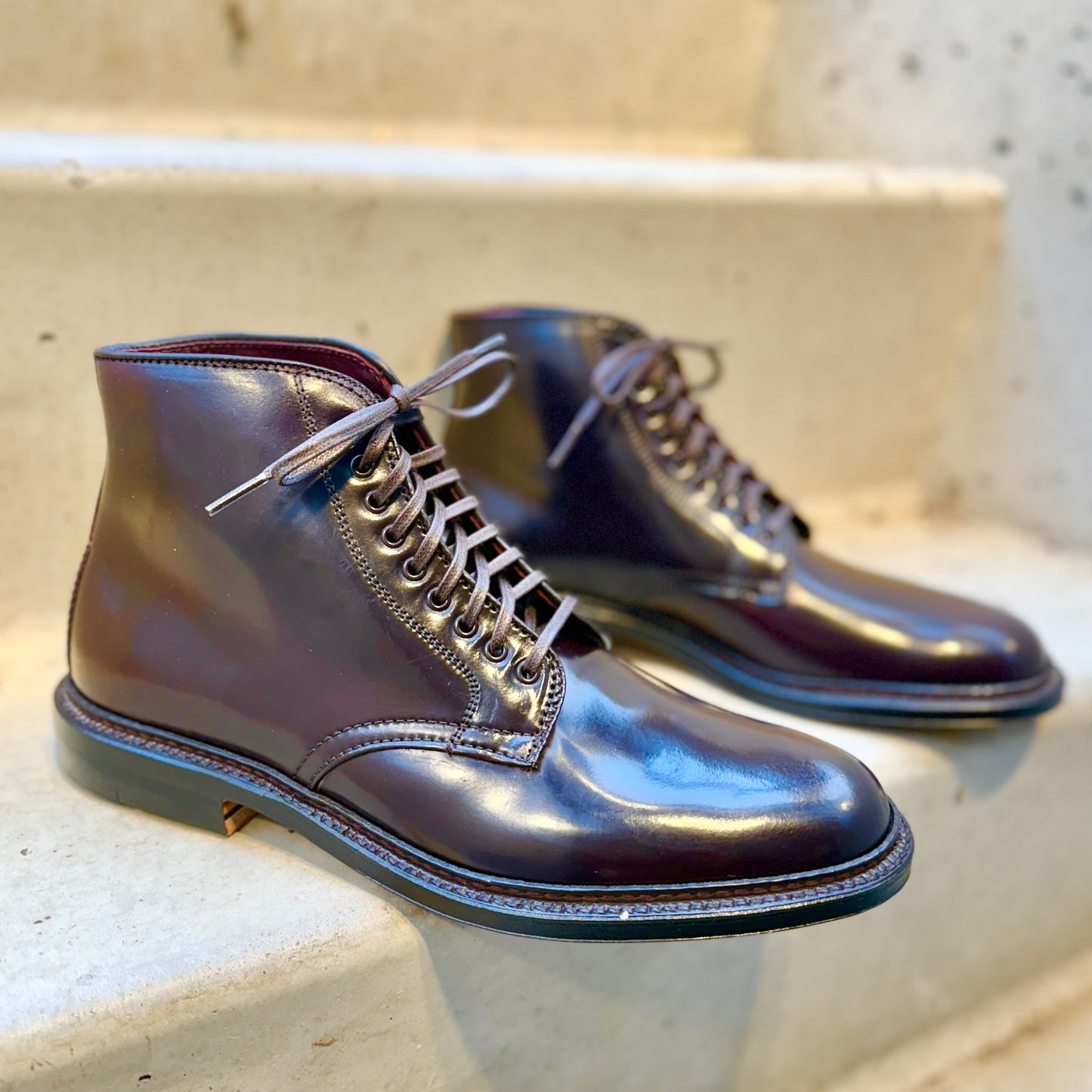 The Alden Madison 45168 - Plain Toe Director Boot in Color 8 Shell Cordovan rests on light-colored concrete steps, featuring sleek polished leather with a glossy finish, laces, and a contrasting Single Oak Leather Sole for a stylish and sophisticated design.