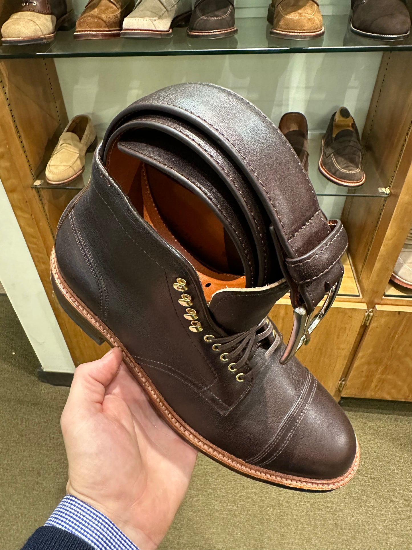 A hand holds a dark brown leather boot with an Alden Madison MB 5334 - Arabica Lux Belt looped through its laces. The boot features brass eyelets, a tan sole, and a nickel buckle. In the background, shoes are showcased in a glass case under soft 35mm wide lighting.
