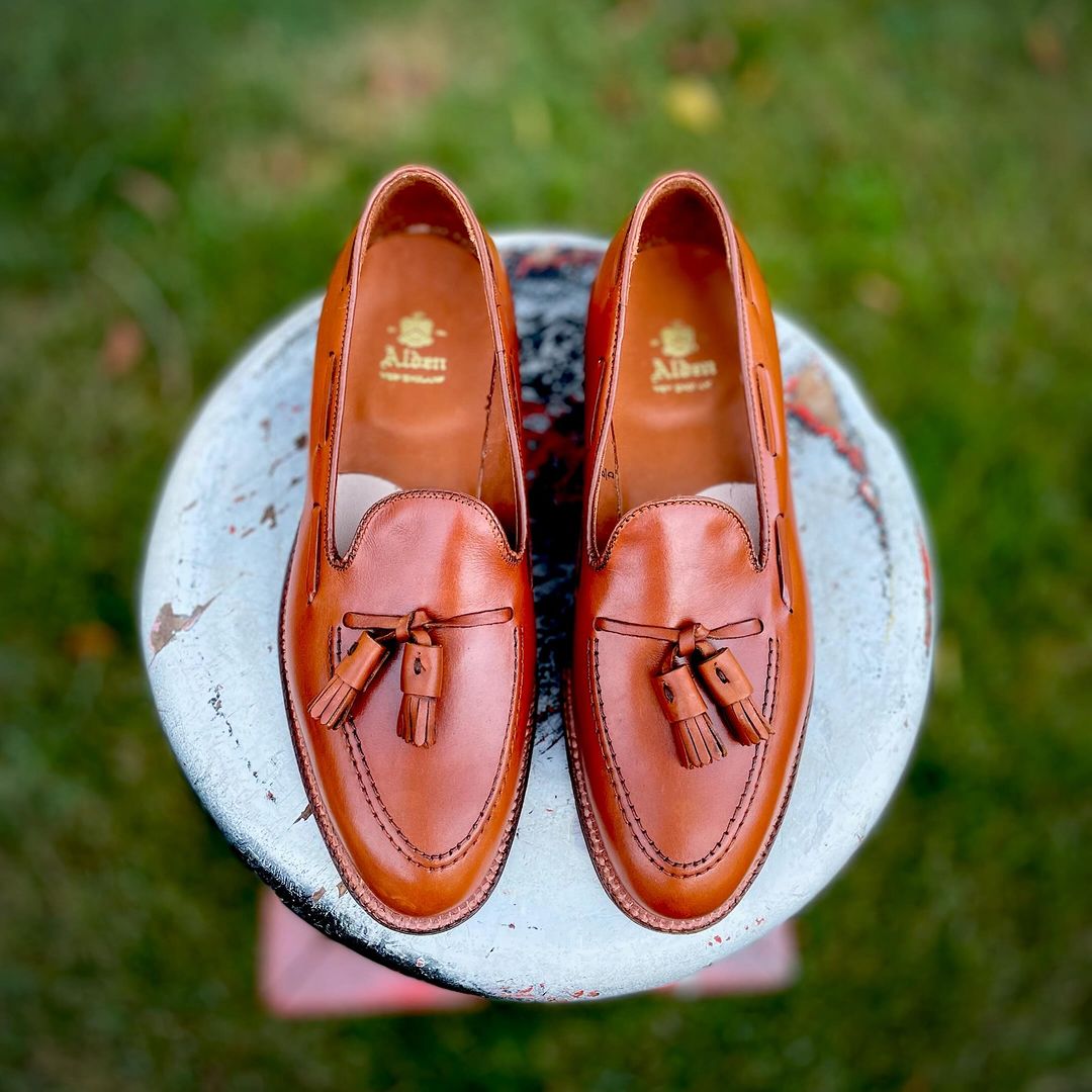 Alden Madisons 662 Tassel Loafers in burnished tan calfskin, featuring decorative stitching and a leather sole on the Aberdeen Last, are elegantly showcased on a weathered round stool outdoors against lush grass.