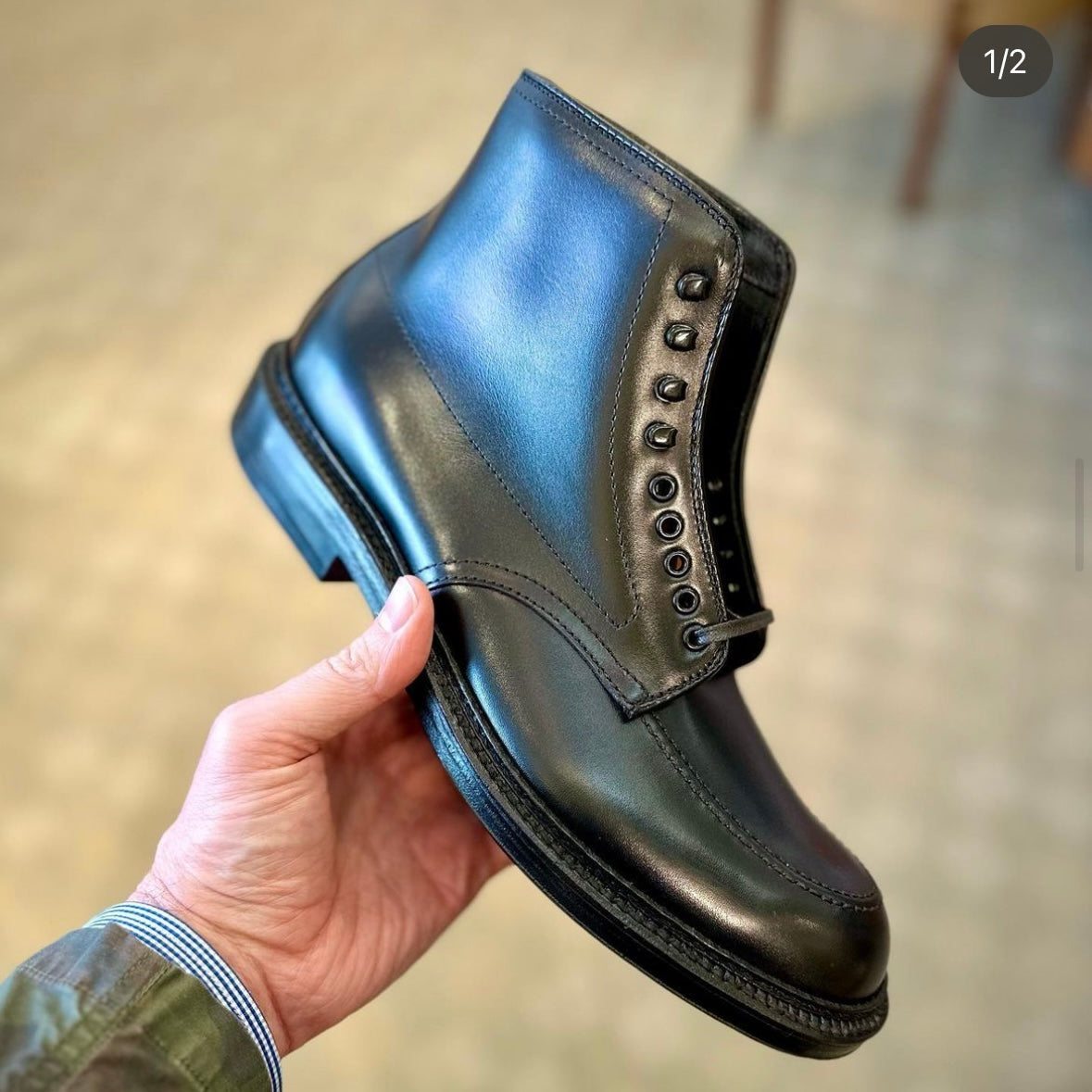 In a hand, the Alden Madison D2941H - Black Calf Indy Boot, dark green in appearance, shines with its multiple eyelets and rounded toe. It features a double leather sole against a blurred tiled floor backdrop.
