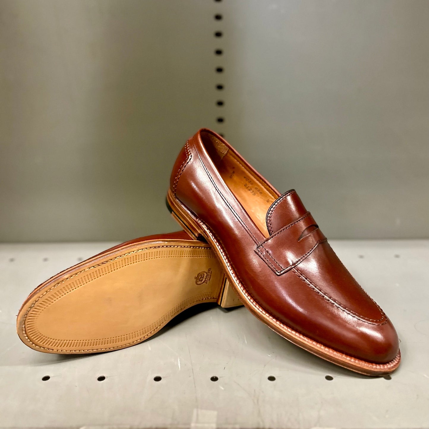 The Alden Madison D1235 - Madison Penny in polished brown calfskin features a smooth upper and stacked heel, with one shoe upright and the other tipped to display its light brown leather sole against a plain gray background.