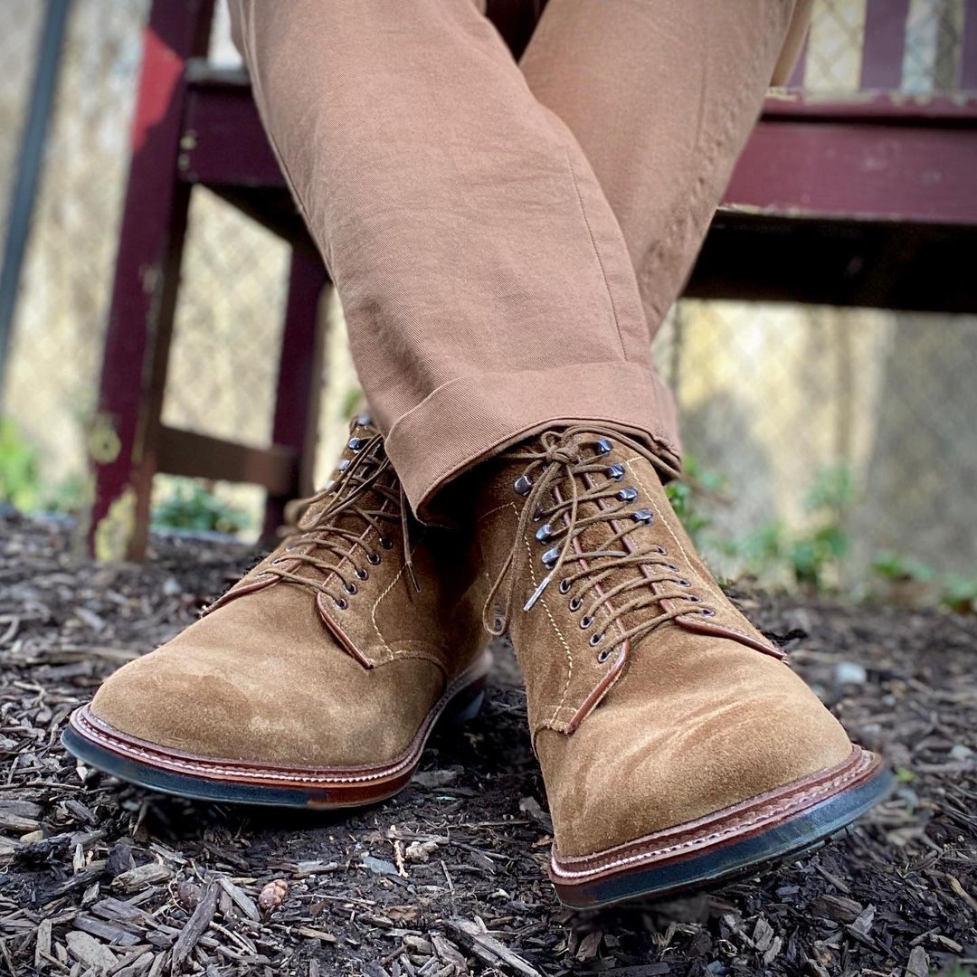 A person sits cross-legged on a bench, wearing Alden Madisons 4511HC Plain Toe Boots in Snuff Suede and brown pants. The background includes a fence, trees, and a wood chip-covered ground.
