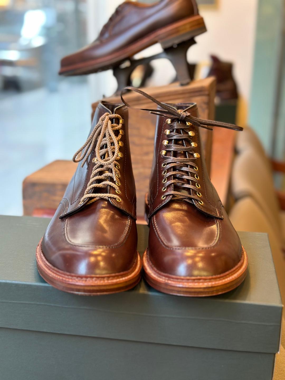 The Alden Madison 40577H - Stitchdown Otis Indy Boot, crafted from luxurious brown calf leather, features contrasting light and dark brown laces and rests on a green box. In the blurred background, another shoe showcases the classic Indy Boot style.