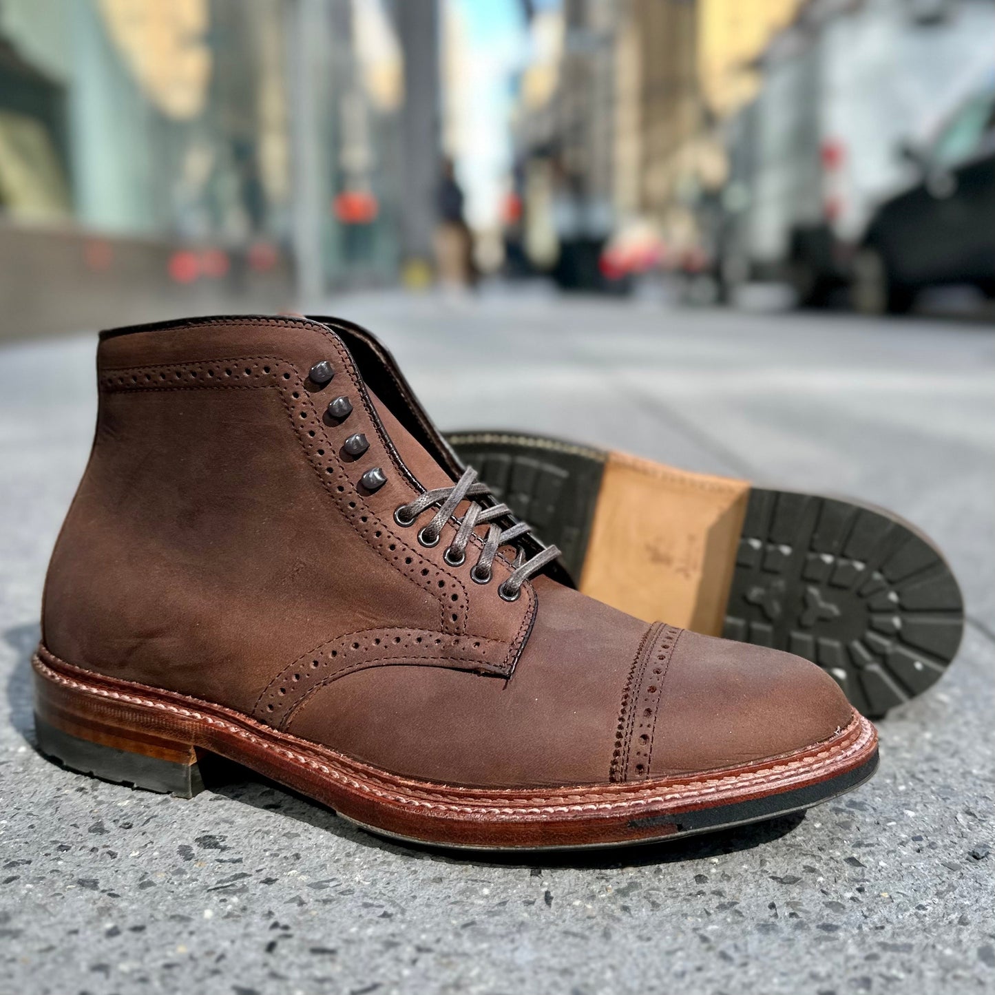A pair of D2817HC - Straight Tip Shaking Boot in Tobacco Smooth Chamois by Alden Madison is on a city street. One boot stands upright and the other lies, displaying its rugged Commando Sole with black eyelets and detailed stitching. Blurred urban buildings are visible in the background.