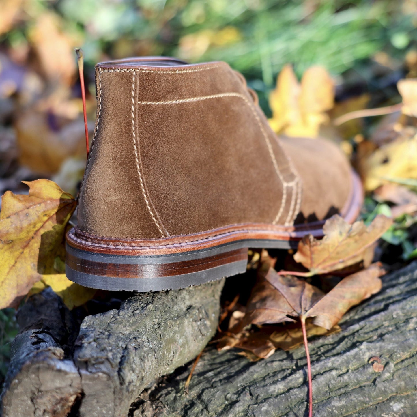 A 1593L Chukka Boot with L3 Sole in Snuff Suede by Alden Madison sits on a log amid autumn leaves, highlighting visible stitching and a slight heel, with the blurred green grass accentuating the Barrie Last craftsmanship.