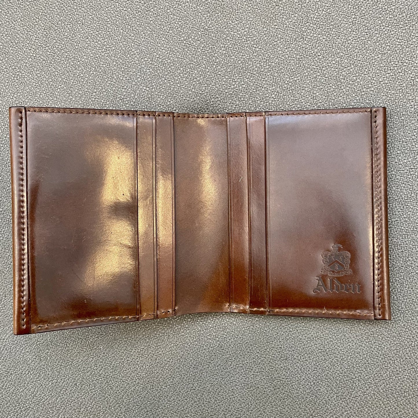 A brown Shell Cordovan wallet, the LG 7007 by Alden Madison, sits on a gray textured surface. With multiple card slots and Alumi embossed on the side, it exudes the classic style of Alden Shoe Co.