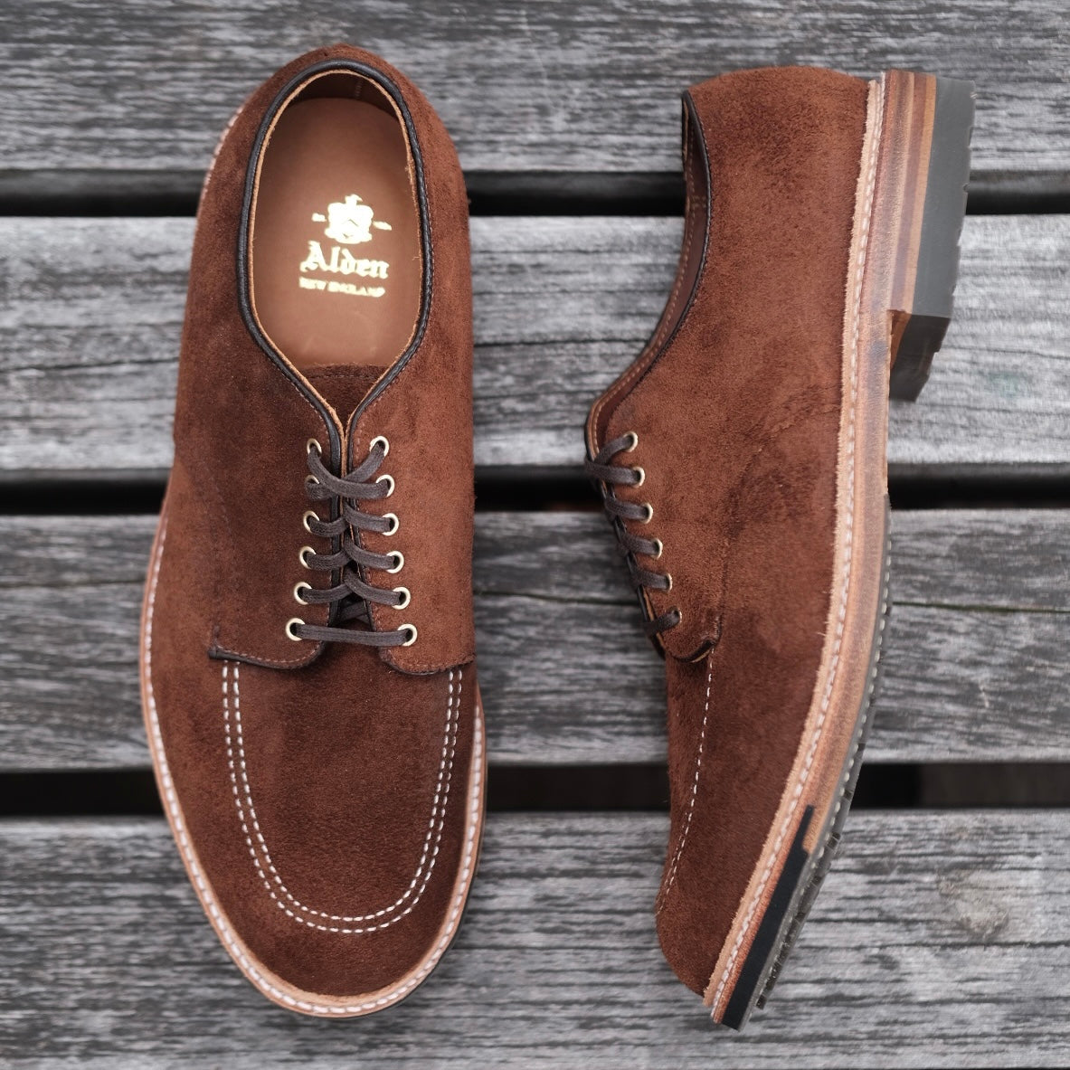 A brown suede D2604C Indy Shoe by Alden Madison sits on a wooden surface. Crafted from Tobacco Reverse Chamois with white stitching, one shoe is upright while the other lies on its side, revealing the sole and Trubalance Last design.