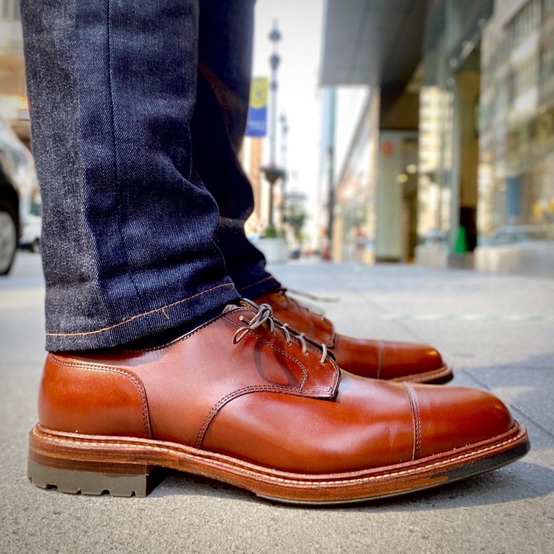 A close-up shows a person wearing Alden Madisons D9539C Ten to One DeRosa Blucher shoes in Madison Brown Calfskin with a Commando Sole, paired with dark blue jeans on a city sidewalk. The background features blurred buildings and pedestrians.