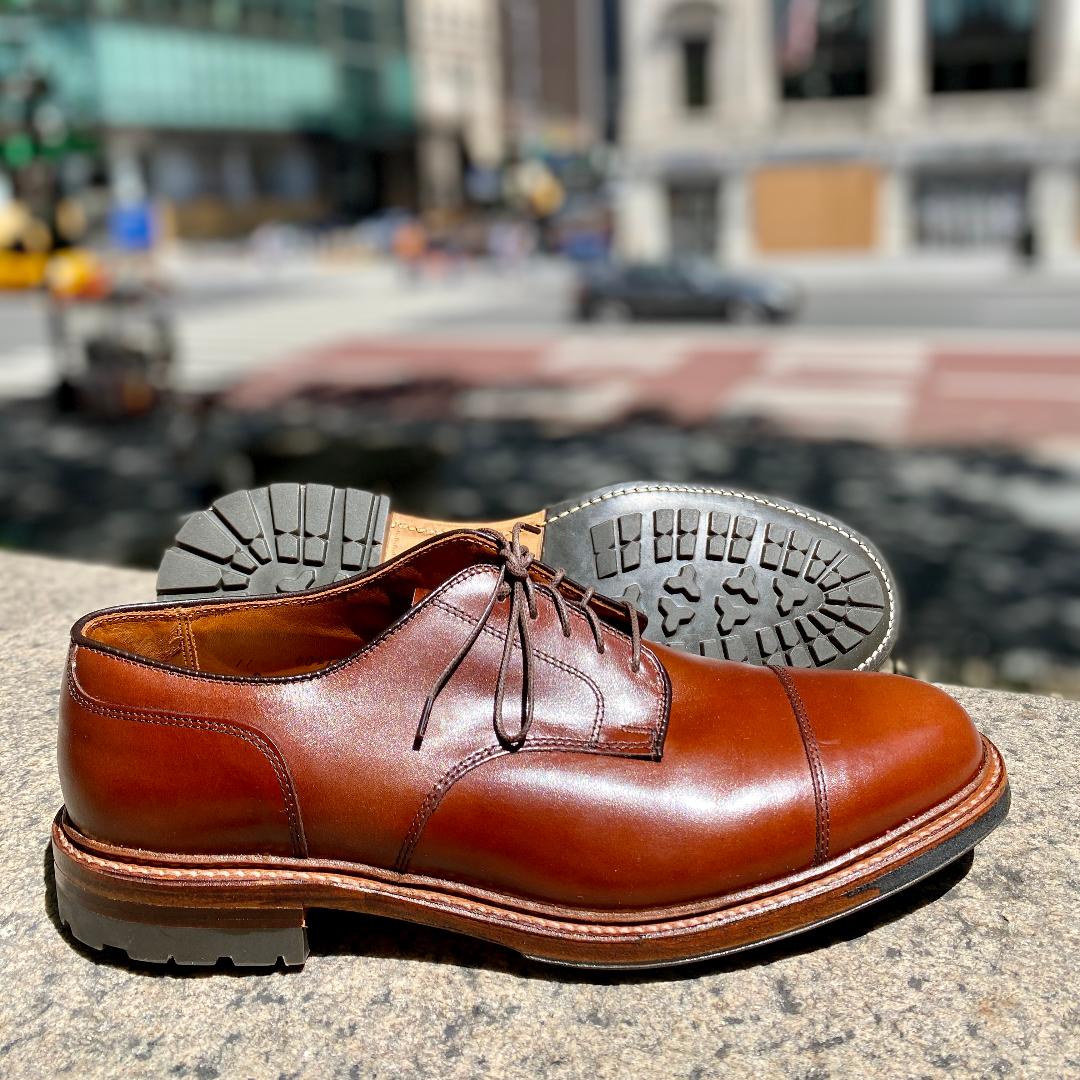 The Ten to One DeRosa Blucher by Alden Madison in Madison Brown Calfskin is displayed outdoors on stone, with one shoe showing its textured Commando Sole. A blurred city street and buildings create a chic backdrop.