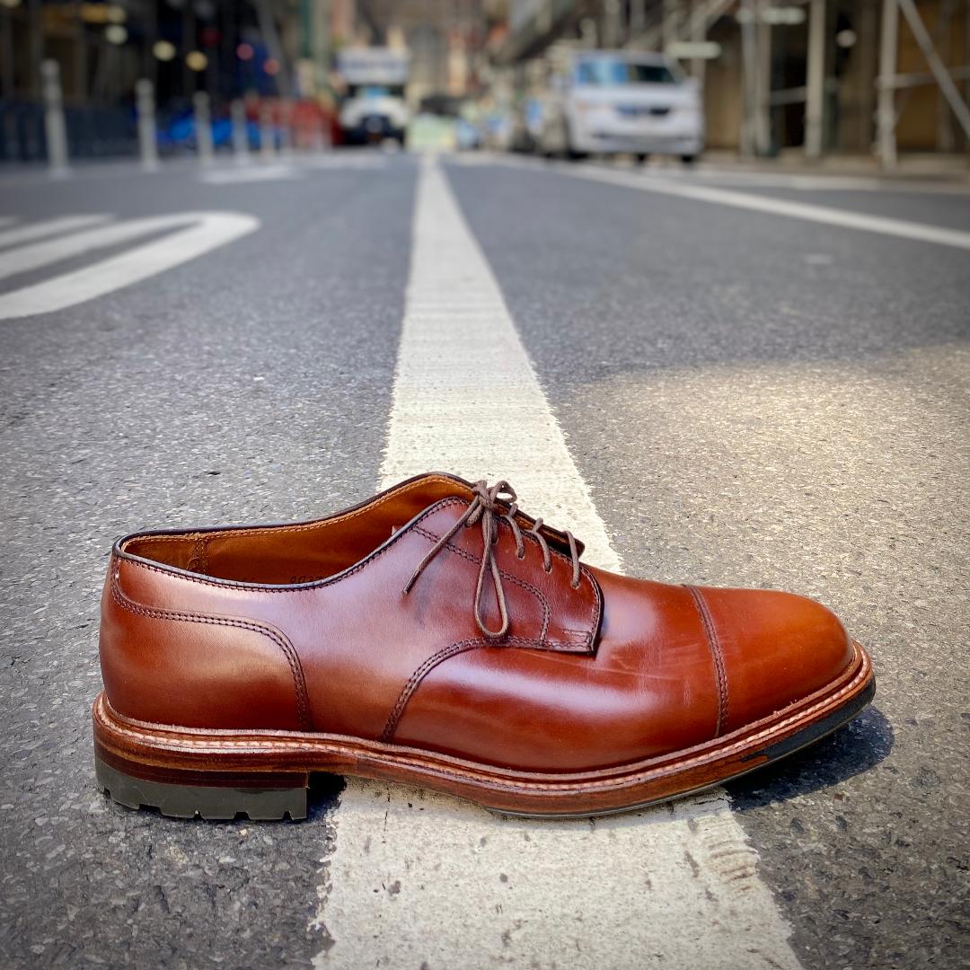 A single D9539C - Ten to One DeRosa Blucher by Alden Madison in brown calfskin, featuring laces and a sleek cap toe, is centered on a white line on an urban street. The background shows blurred buildings and vehicles.