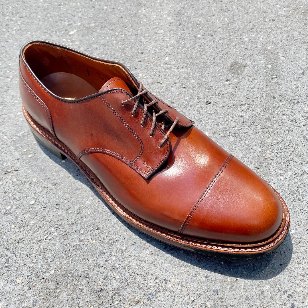 A DeRosa Blucher by Alden Madison, Ten to One (D9539C) in Madison Brown Calfskin, with laces and a Commando Sole, displays its smooth finish and cap toe design on a textured gray surface under bright sunlight.