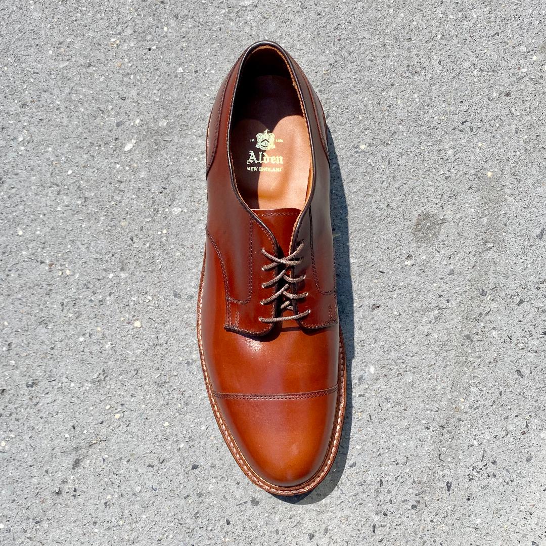 The D9539C Ten to One DeRosa Blucher by Alden Madison in Madison Brown Calfskin, featuring dark brown laces and a Commando Sole, rests on a light gray concrete surface. Captured from above, its polished finish and exquisite stitching details are highlighted.