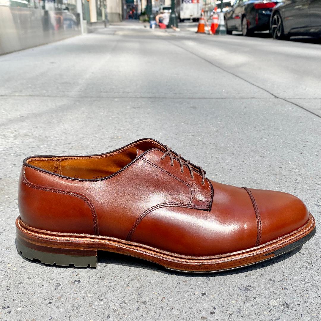 A single Alden Madison D9539C Ten to One DeRosa Blucher in Madison Brown Calfskin, with a cap toe design, laces, and a sturdy Commando Sole, sits on a city sidewalk amid cars and buildings.