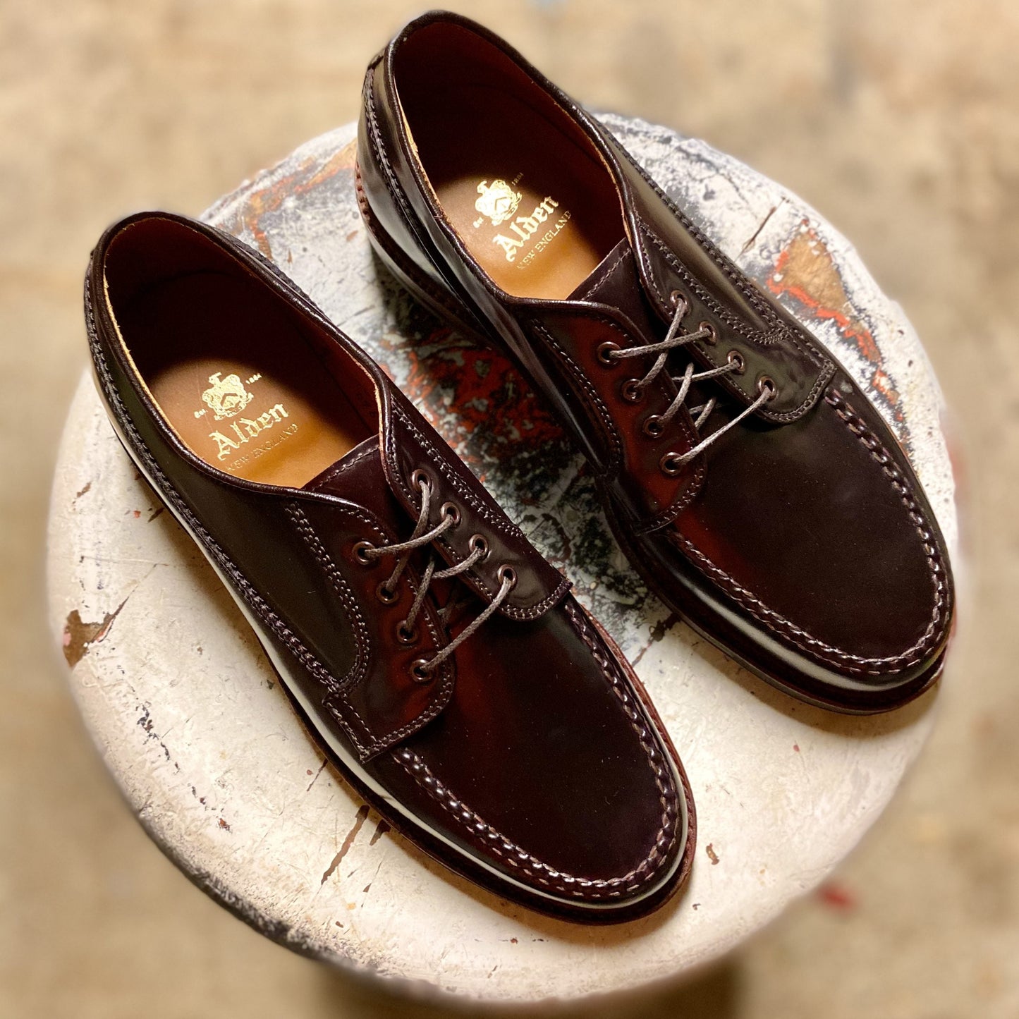A pair of Alden Madison D9613C Ranger Mocc shoes in Color 8 Shell Cordovan, featuring polished leather, visible stitching, brown laces, and a flat welt, sit on a round, weathered white stool.