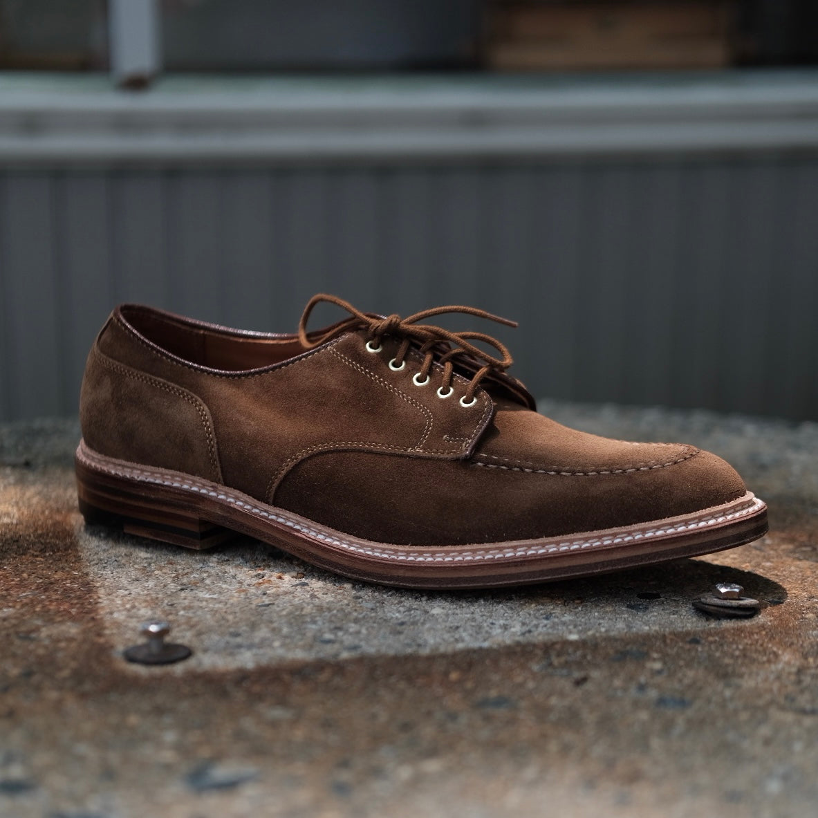 The Quickley in Snuff Suede by Alden Madison is displayed as a single brown dress shoe with laces on a textured surface. With a wooden sole and white stitching, this U Tip Blucher elegantly stands out against a blurred background.