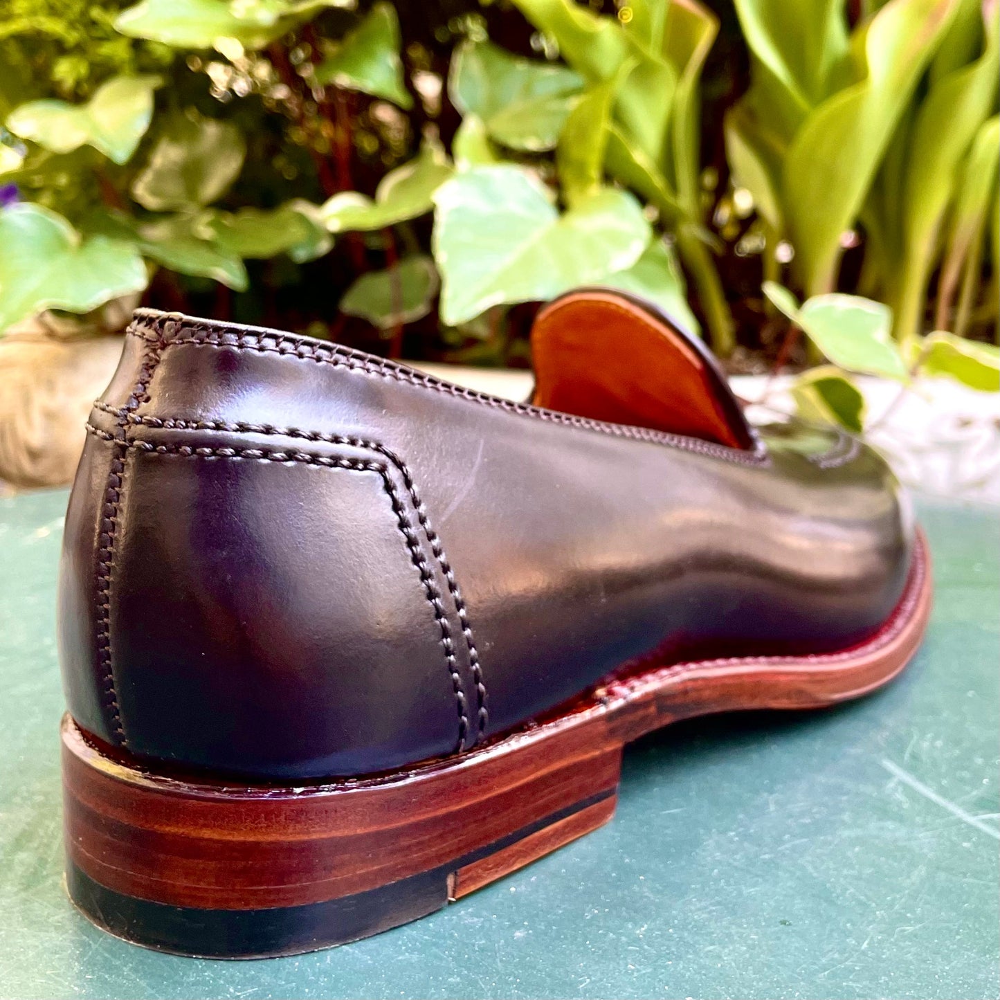 A close-up of a single Alden Madison D1118 - Vanderbilt Slip On in Color 8 Shell Cordovan, crafted with visible stitching, rests on a green surface. Displayed on the Aberdeen Last, the shoe is positioned heel-forward against a backdrop of lush green leaves.