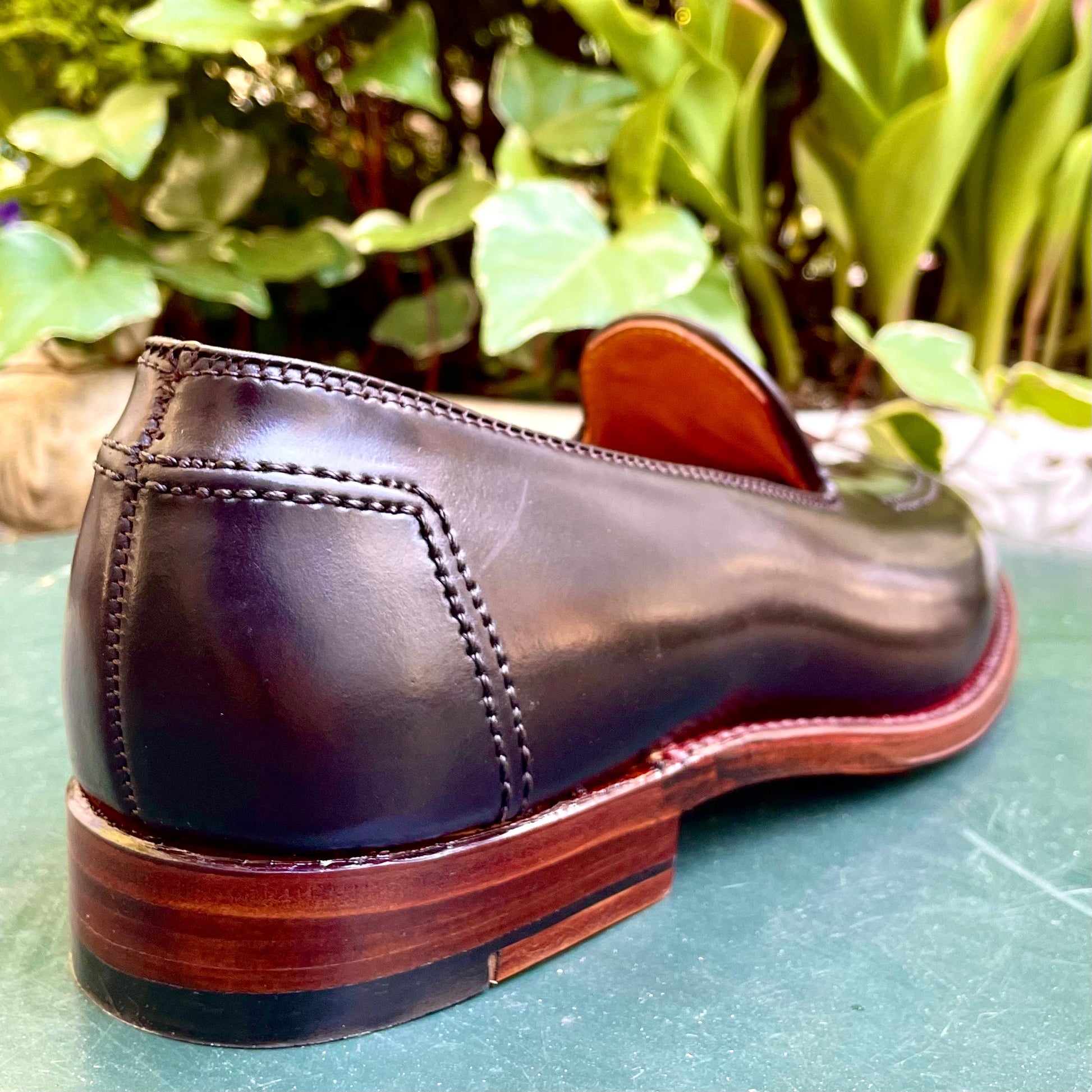 A close-up of a single Alden Madison D1118 - Vanderbilt Slip On in Color 8 Shell Cordovan, crafted with visible stitching, rests on a green surface. Displayed on the Aberdeen Last, the shoe is positioned heel-forward against a backdrop of lush green leaves.