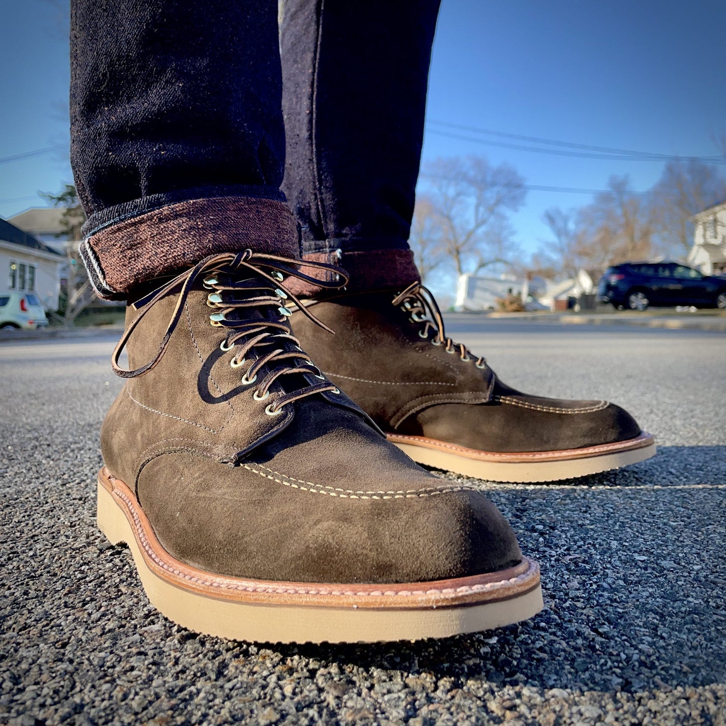On a clear day, someone stands on the pavement wearing Alden Madisons D0938H - Duncan Indy Boot in Loden Suede with light soles, their jeans rolled to reveal socks, against a backdrop of a street and trees.