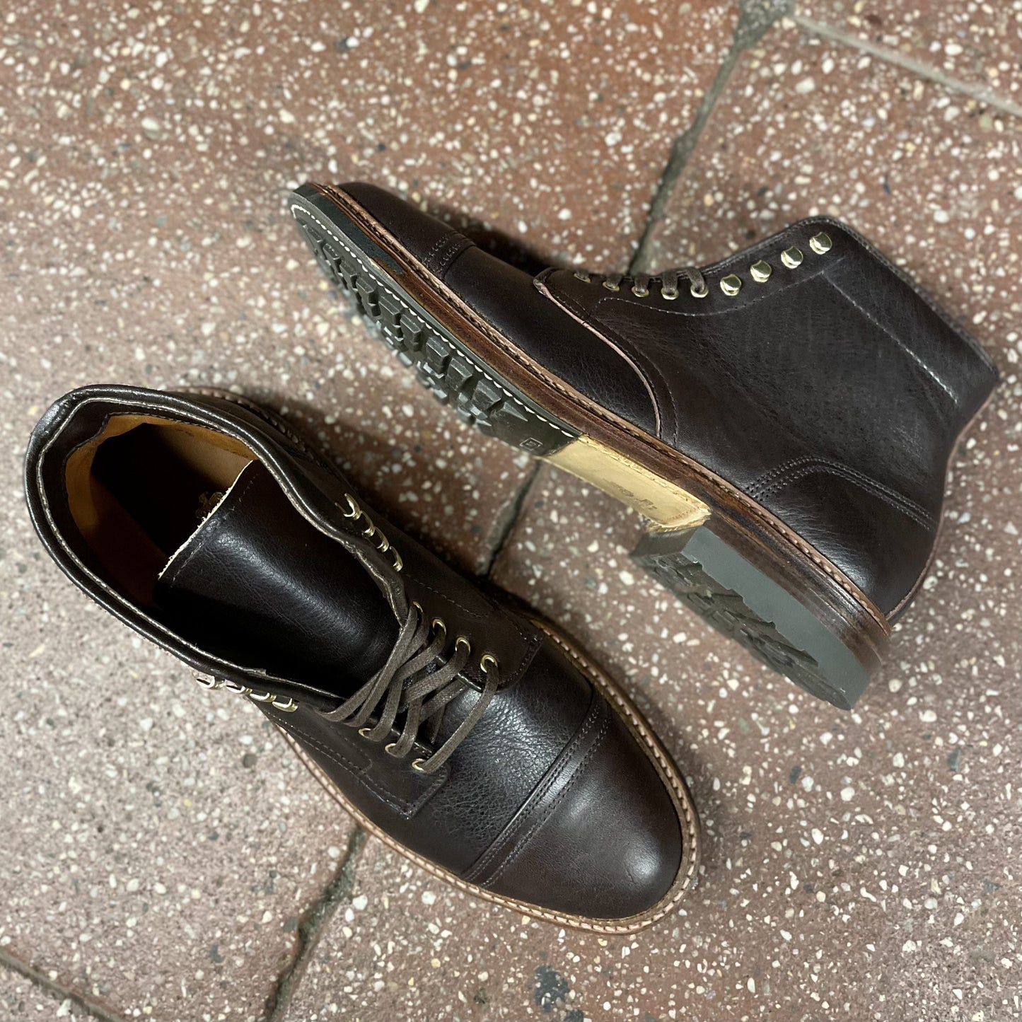 A pair of Alden Madison D1808HC - Stitchup 2/2 Boots in dark brown Arabica Lux7 calfskin with laces and gold eyelets, featuring a Commando sole, rest on a speckled reddish-brown tile—one upright, one on its side showing the rugged sole.