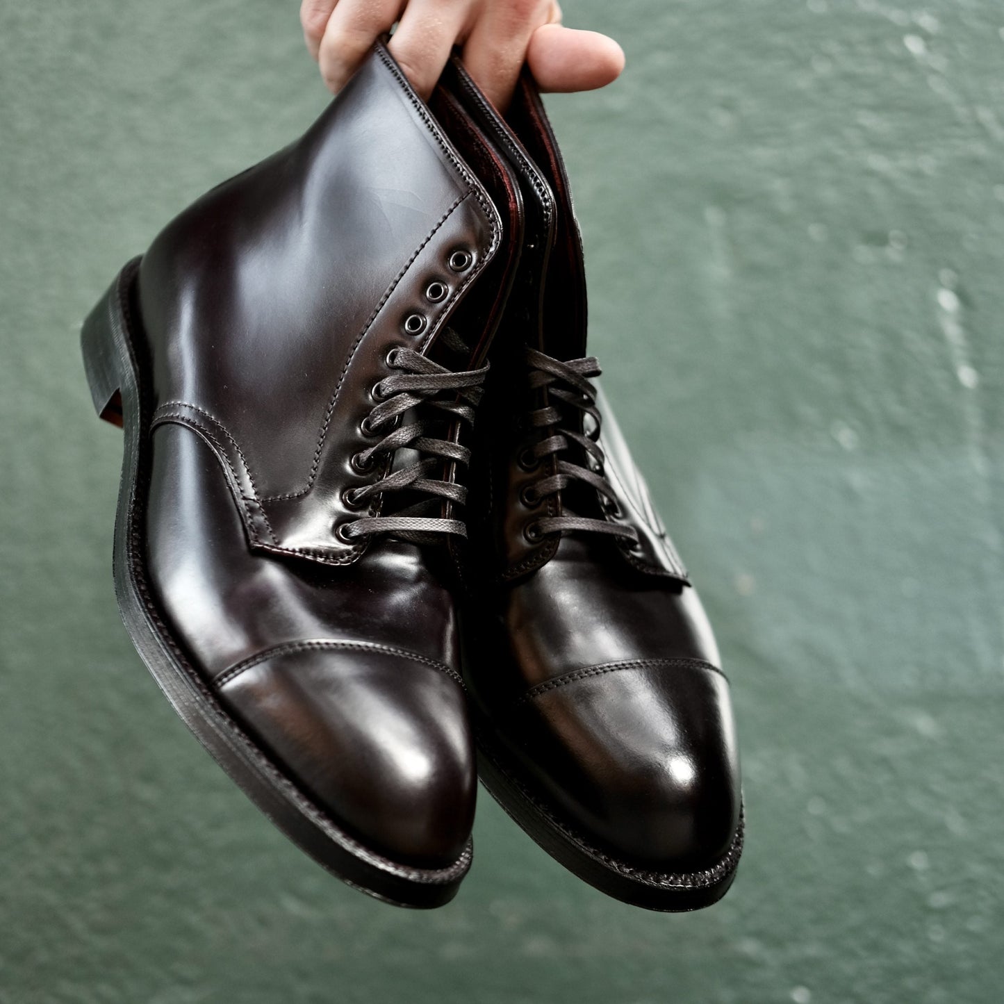 A hand holds Alden Madisons 4060 Cap Toe Boots in polished black Shell Cordovan, featuring a Double Oak Leather Sole, against a textured green background. The boots look new and shiny with their elegant design.