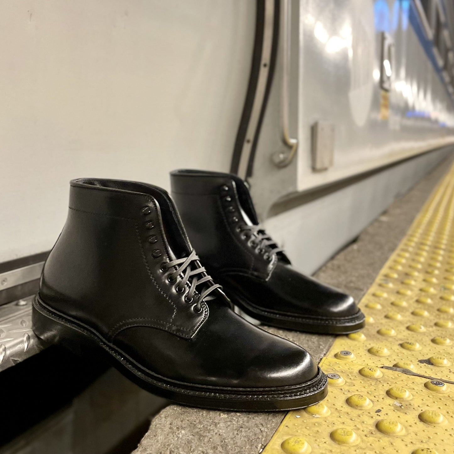 On a concrete subway platform near a yellow tactile paving strip sits the Alden Madison D6842H Plain Toe Director Boot in Black Shell Cordovan, featuring speed hooks. The blurred background showcases a stationary train with metal doors and a white exterior.