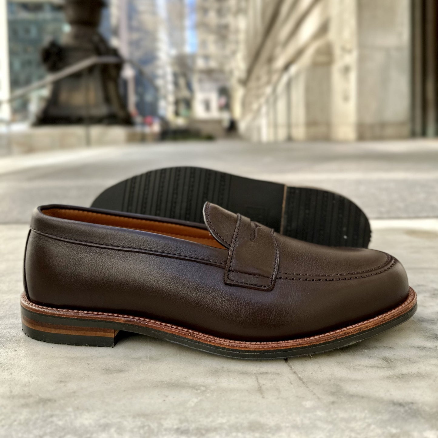An Alden Madison D2203L Penny Loafer in Brown Lady Calf with an Antique Flat Welt rests on a marble surface outdoors, its stitched sole elegantly highlighted against a softly blurred urban background with buildings and a lamppost.