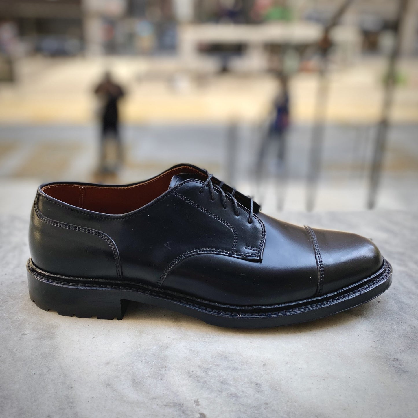 The Alden Madison 2175C - DeRosa Captoe Blucher in Black Shell sits on a gray concrete surface, showcasing clean stitching and a glossy finish. Its sleek Commando Sole is perfect for urban adventures against a blurred background with muted urban elements and people.