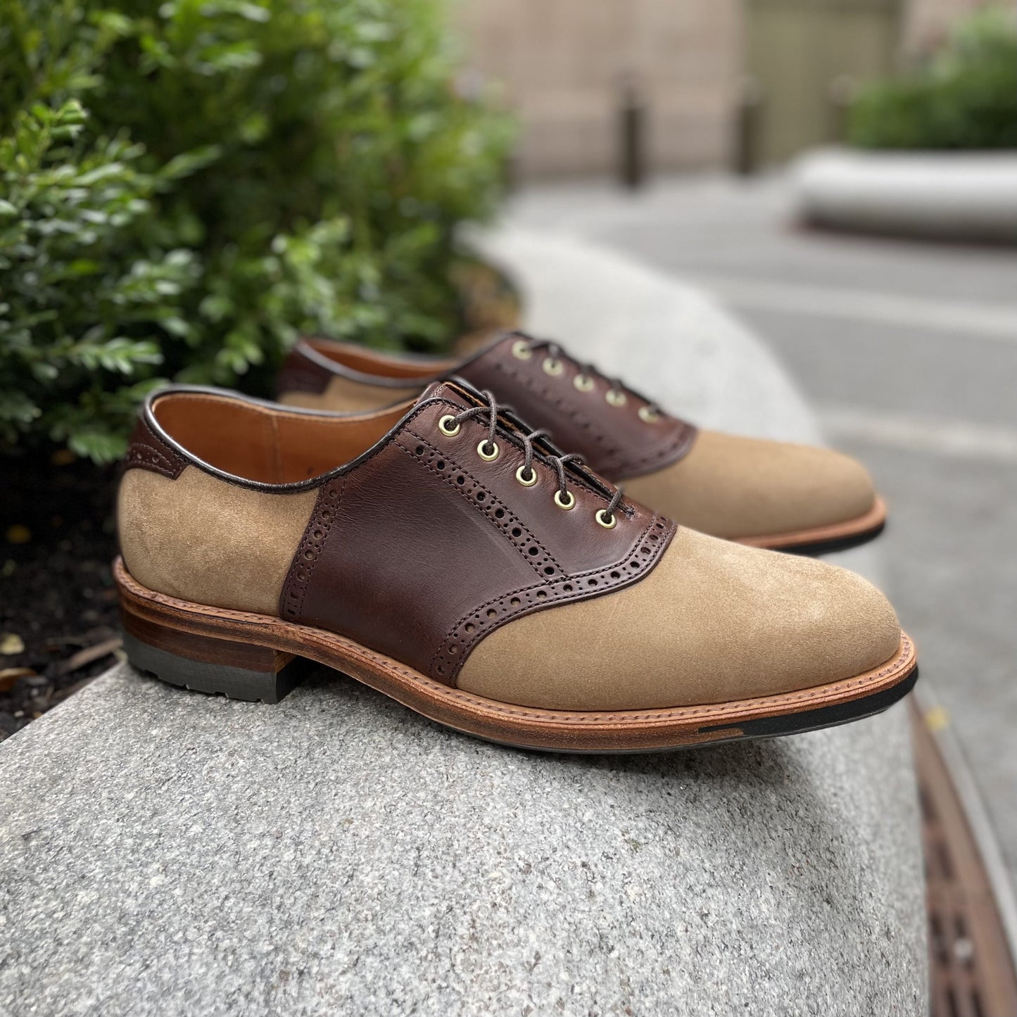 Aldem Madisons D1304C - Valhalla Saddle Oxford in CXL and Tan Suede, with a Commando Sole, rests stylishly on a smooth, curved concrete surface. Lush greenery, blurred in the background, suggests an outdoor setting.