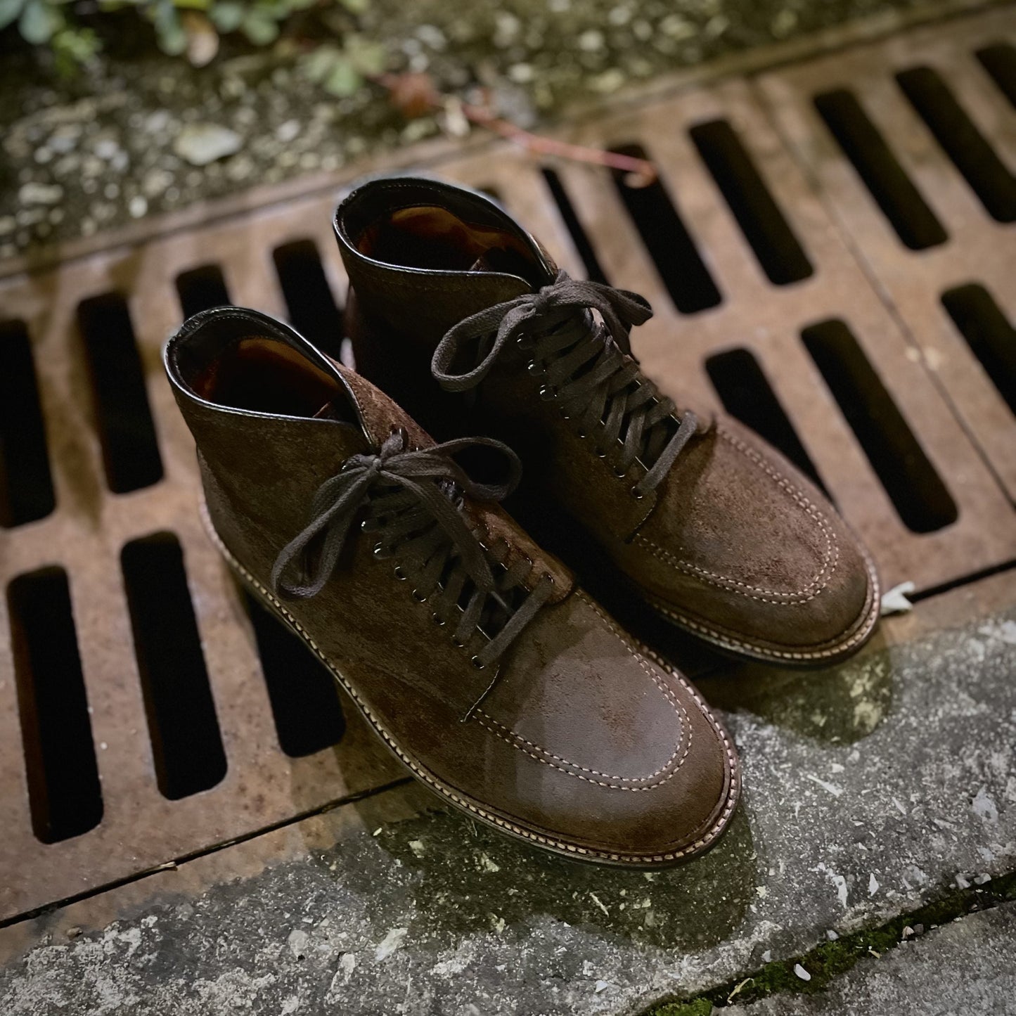 A pair of 4015HC - Indy Boot in Tobacco Reverse Chamois by Alden Madison, featuring visible stitching and Commando Soles, rests on a damp surface by a metal grate. With their slightly worn appearance, they sit amid faint green foliage in the background.