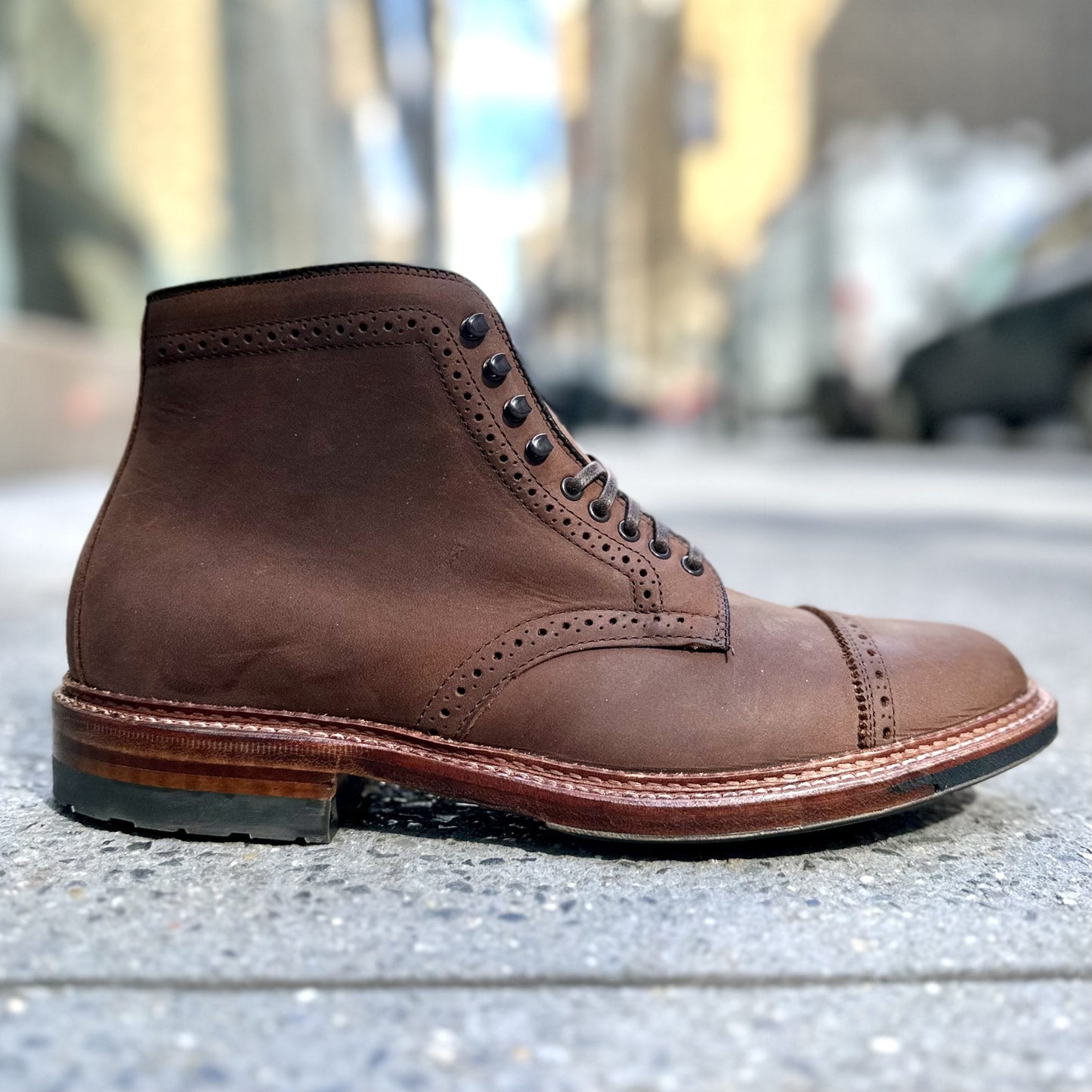 A single D2817HC - Straight Tip Shaking Boot in Tobacco Smooth Chamois by Alden Madison, featuring detailed stitching and a wooden sole, is displayed on a city sidewalk. The blurred background shows buildings and cars under a clear sky.