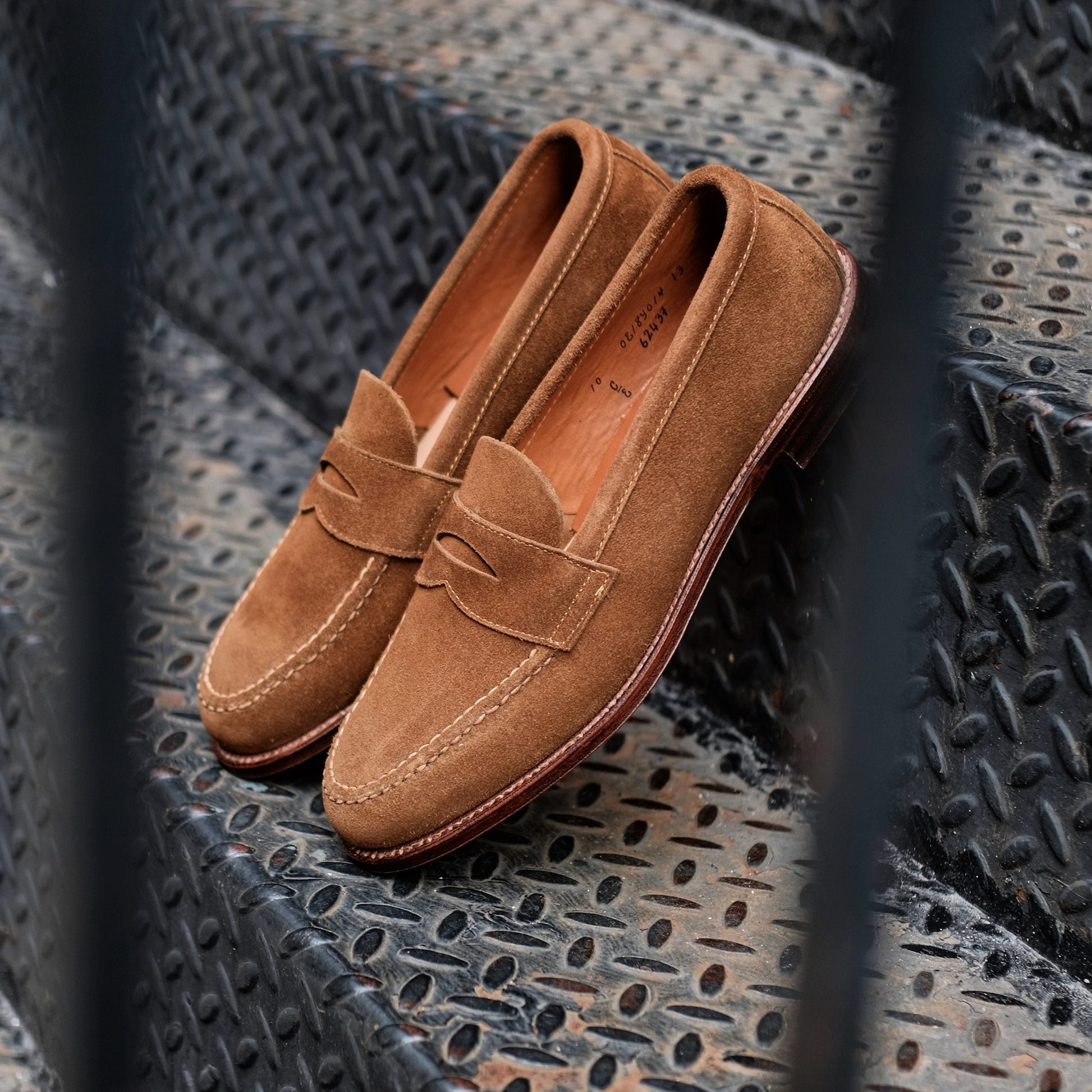 The Alden Madison 6243F - Unlined Leisure Hand Sewn in Snuff Suede loafers, resting on a textured metal stairway, showcase classic penny loafer style with meticulous hand stitching.