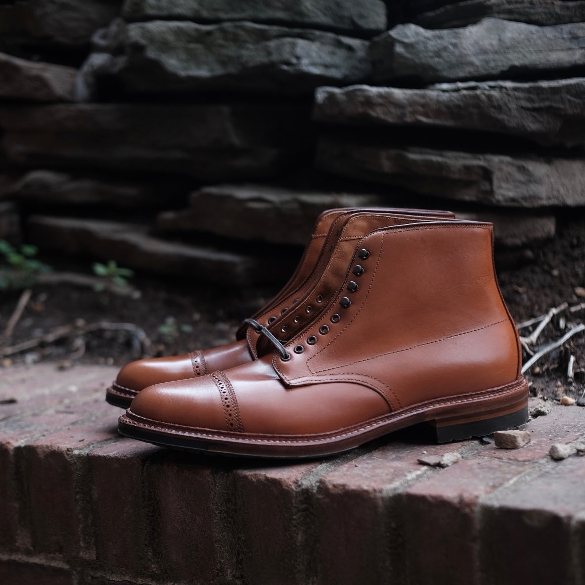 The Alden Madison D2825HC Jumper Boot in burnished tan calfskin rests on a brick surface, surrounded by stacked stones and greenery.