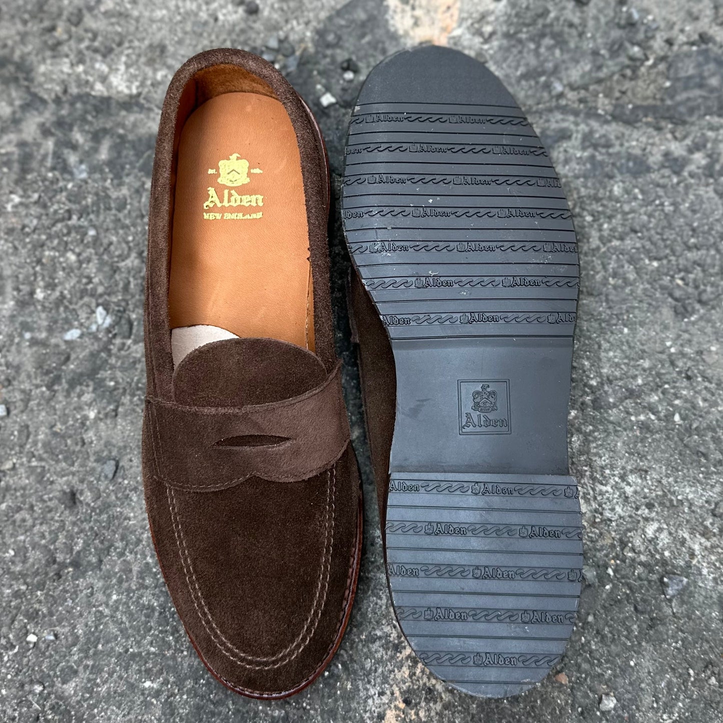 Brown suede Alden Madison 6224L - Unlined Penny Mocc loafers with a moc-toe design rest elegantly on a rough gray surface. The left shoes insole features the brand logo, while the right one displays its textured rubber sole with distinctive Alden branding.