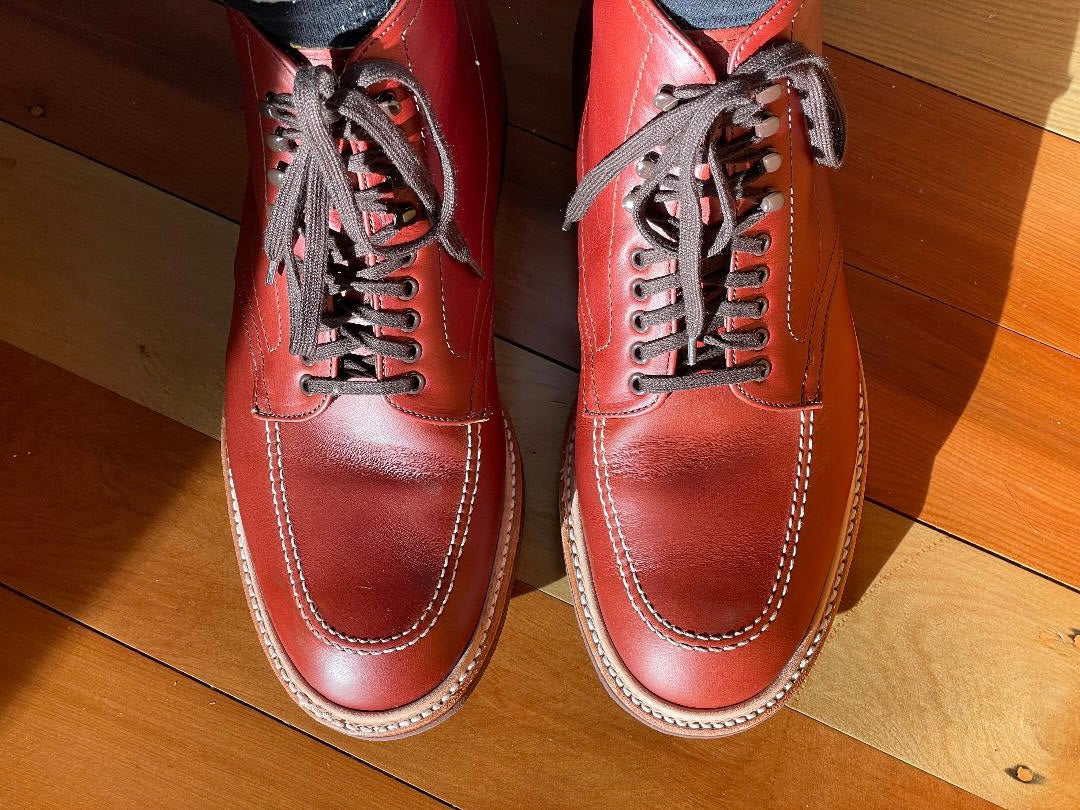 The Alden Madison 405 - Indy Boot in Mid Brown Calfskin, featuring polished leather and dark laces, is showcased from above on a wooden floor. The sunlight highlights the shoes sheen and enhances the rich wood tones.