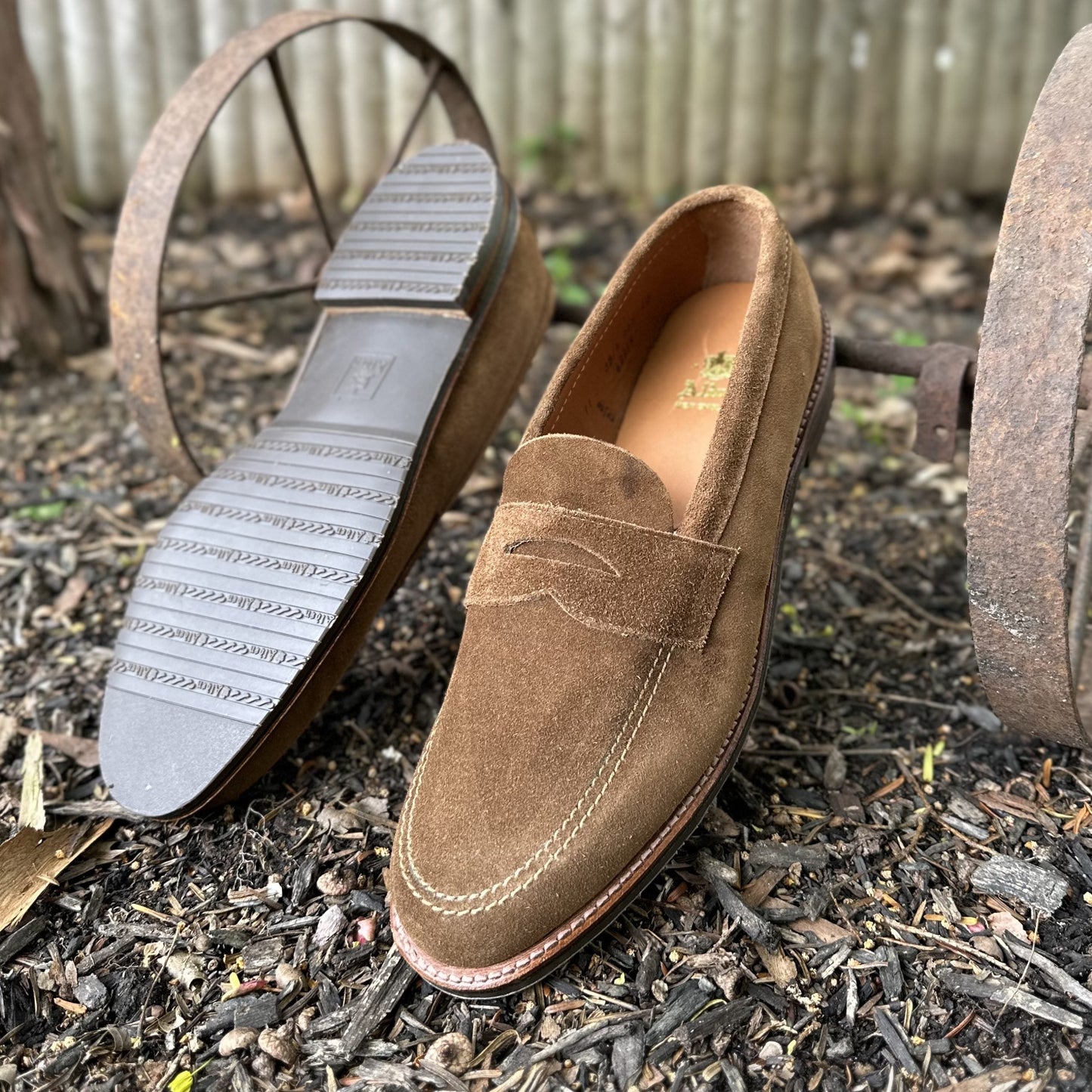 The 6221L - Unlined Penny Mocc in Snuff Suede from Alden Madison, featuring a rustic moc toe design, rests on wood chips. One shoe stands upright as the other reveals its textured rubber sole against a backdrop of an artfully blurred old, rusty metal wheel.