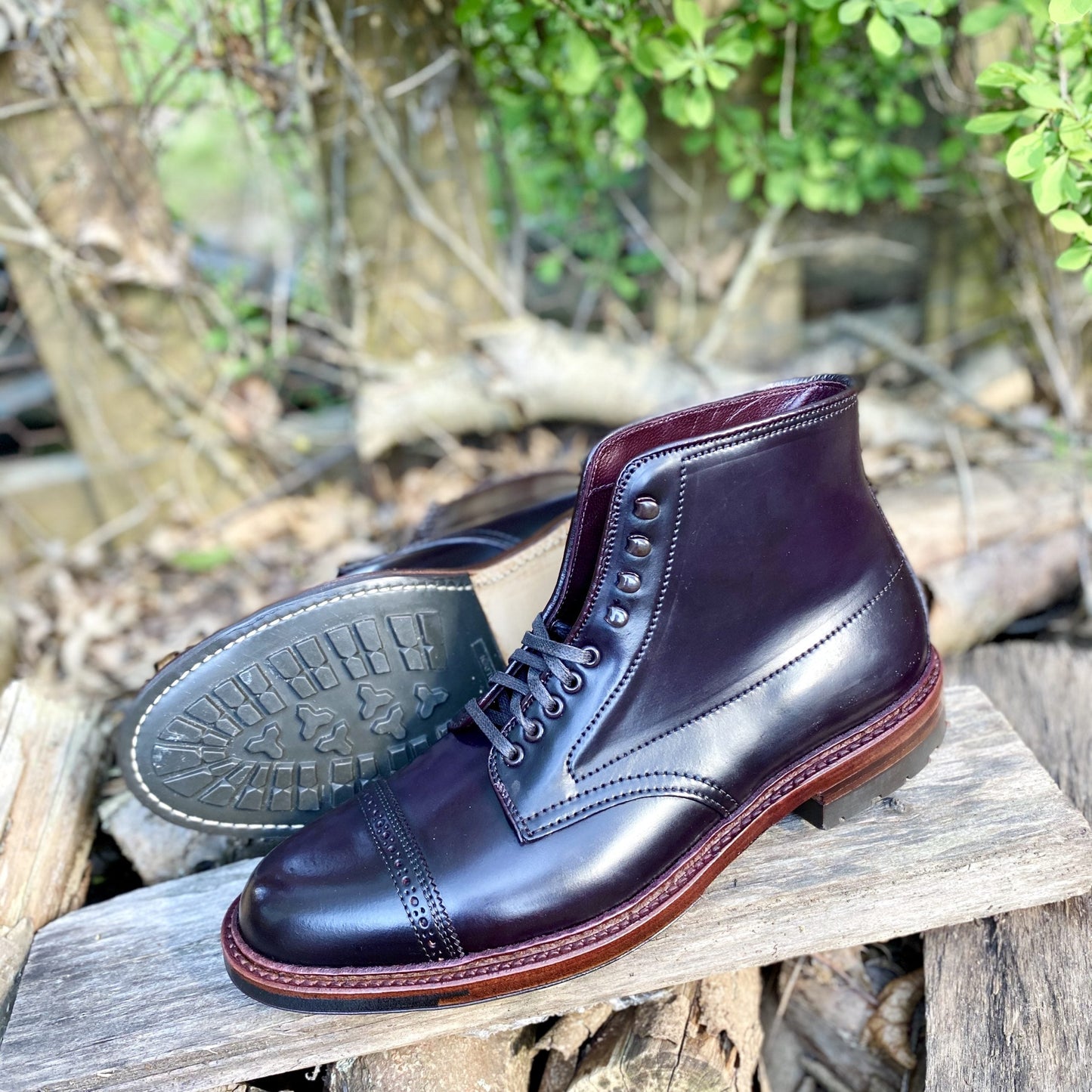 The Alden Madison D6861HC Jumper Boot in Color 8 Shell Cordovan, featuring polished cap toes, Speed Hook Lacing, silver eyelets, and a textured rubber sole, is showcased outdoors on wooden logs with greenery and fencing behind.