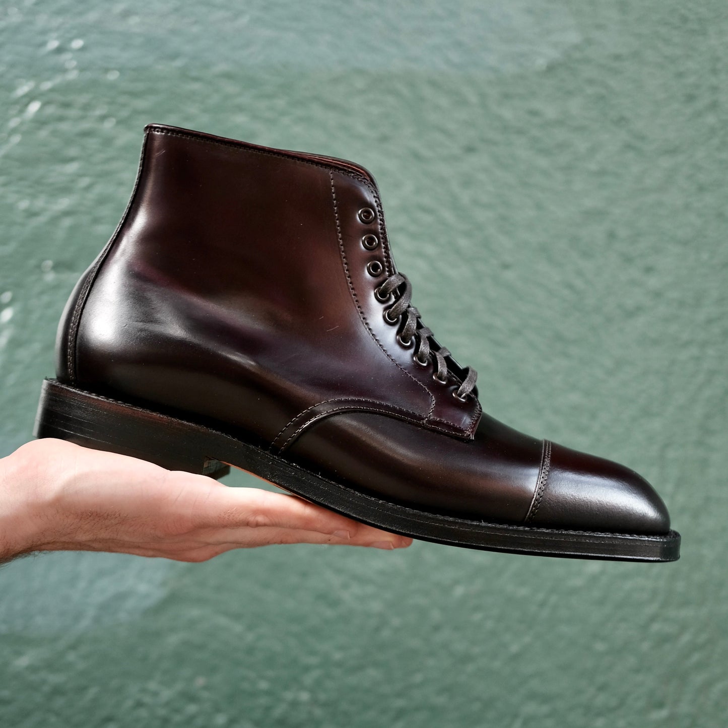 The Alden Madison 4060 Cap Toe Boot in Color 8 Shell Cordovan, a polished dark brown leather boot with laces crafted on the Grant Last, is showcased in a hand against a textured green background.