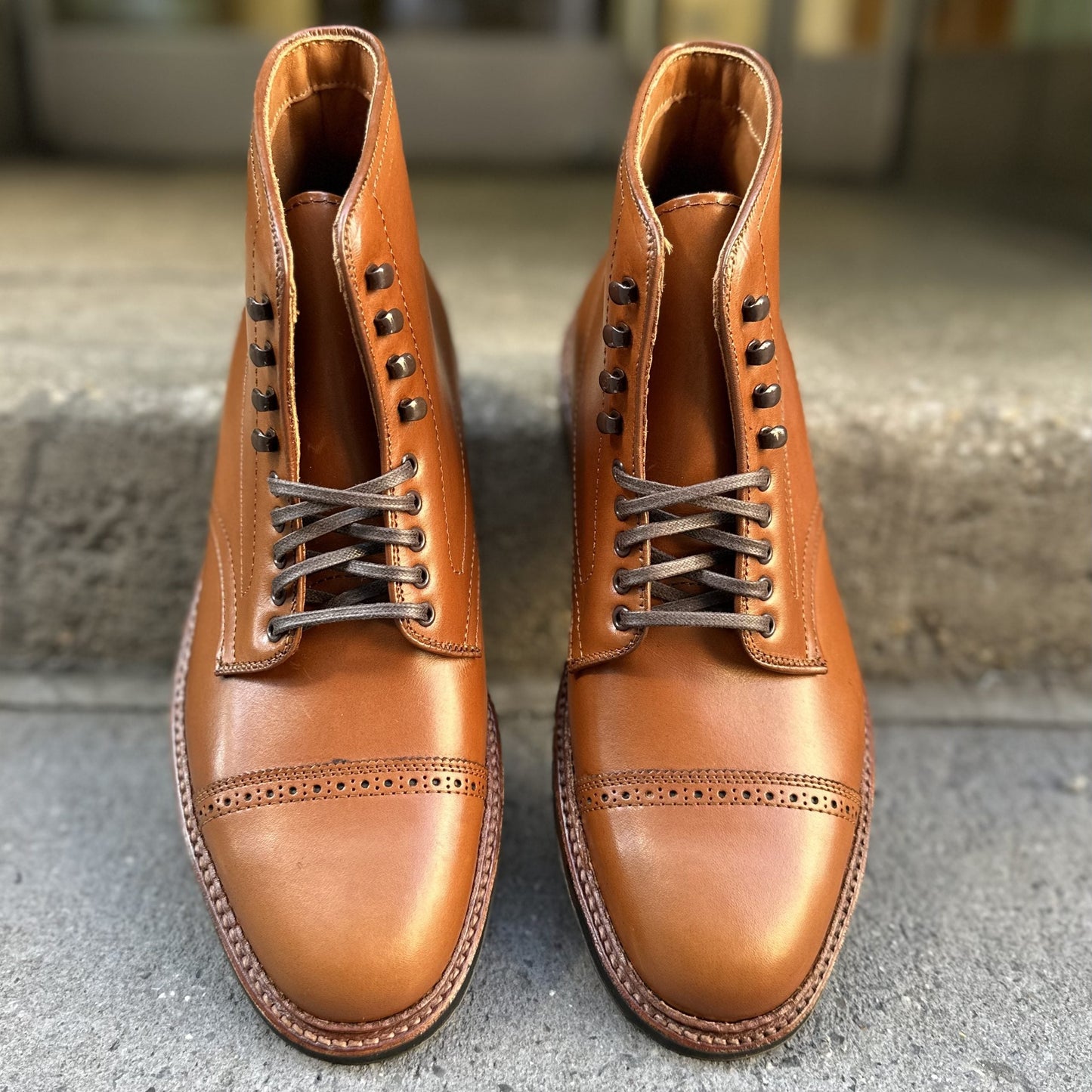 Displayed on a concrete surface, the Alden Madison D2825HC Jumper Boots in burnished tan calfskin feature polished brown leather with laces, brogue detailing on the toe caps, and classic Barrie Last construction, showcasing their design and artistry.