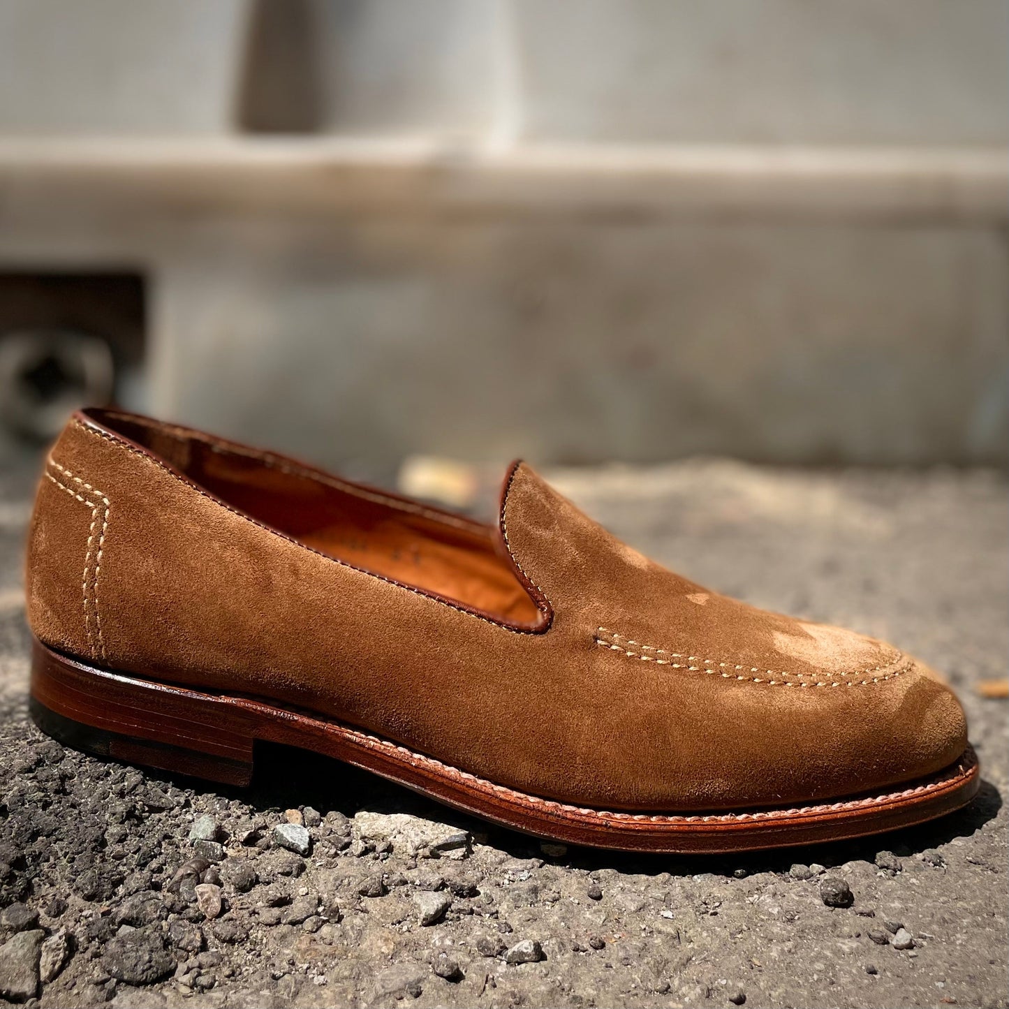 A D2101 - Vanderbilt Slip On in snuff suede by Alden Madison rests on a textured, pebbled surface, showcasing its visible stitching and a wooden Brooks heel against a softly blurred background.