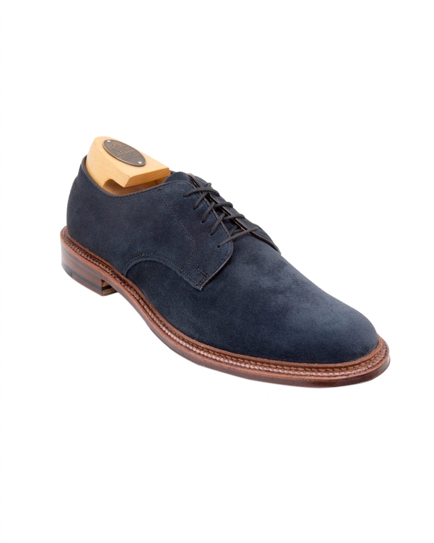 The Alden Madison 29331F - Unlined Plain Toe Blucher in blue suede, with black laces and a brown flex leather sole, is showcased on a white background. A wooden shoe tree retains its shape, complemented by the expert storm welt stitching for added craftsmanship.