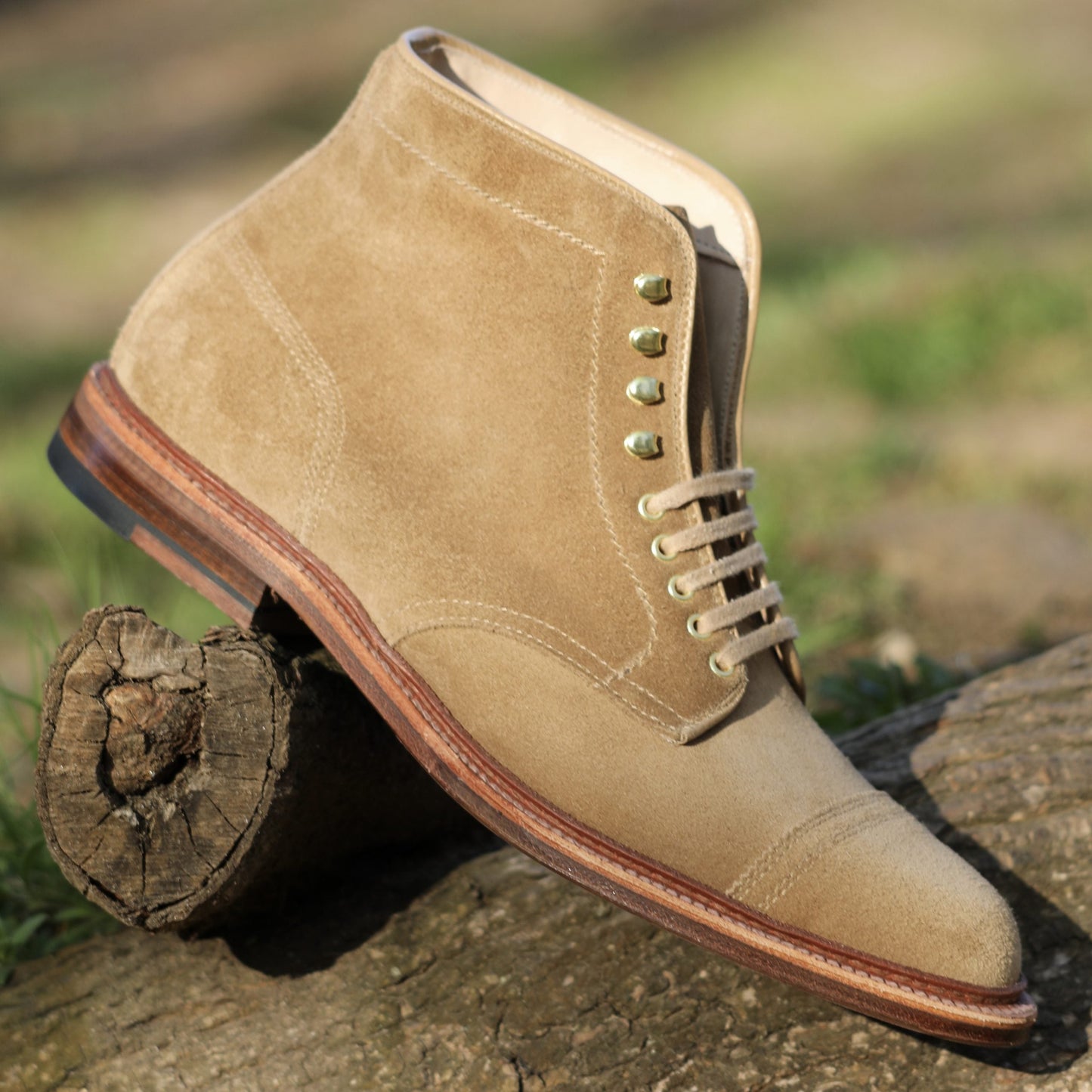 The D2685H - 2/2 Cap Toe Boot in tan suede by Alden Madison features brass eyelets, a smooth toe cap, and a brown leather sole. Elegantly placed on a log outdoors with neatly tied laces, its rugged charm is enhanced by the blurred grassy background.