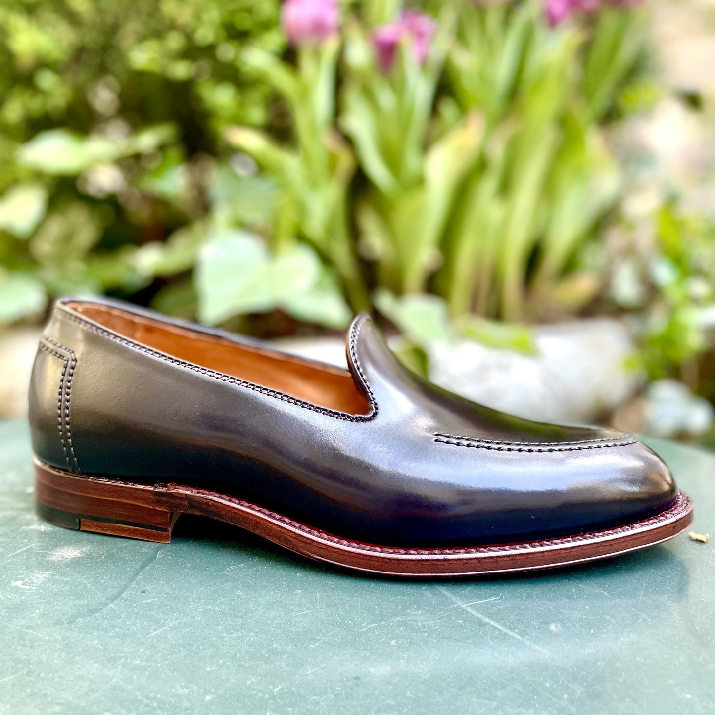 The Alden Madison D1118 - Vanderbilt Slip On in Color 8 Shell, a polished leather loafer with a wooden sole and brown stitching, is displayed on a green surface. In the blurred background, greenery blends with purple flowers.