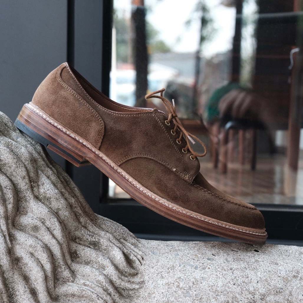 The 24082 - The Quickley in Snuff Suede by Alden Madison, featuring a leather sole, is elegantly placed on a textured stone surface, illuminated by soft window light that reflects a blurred indoor scene.