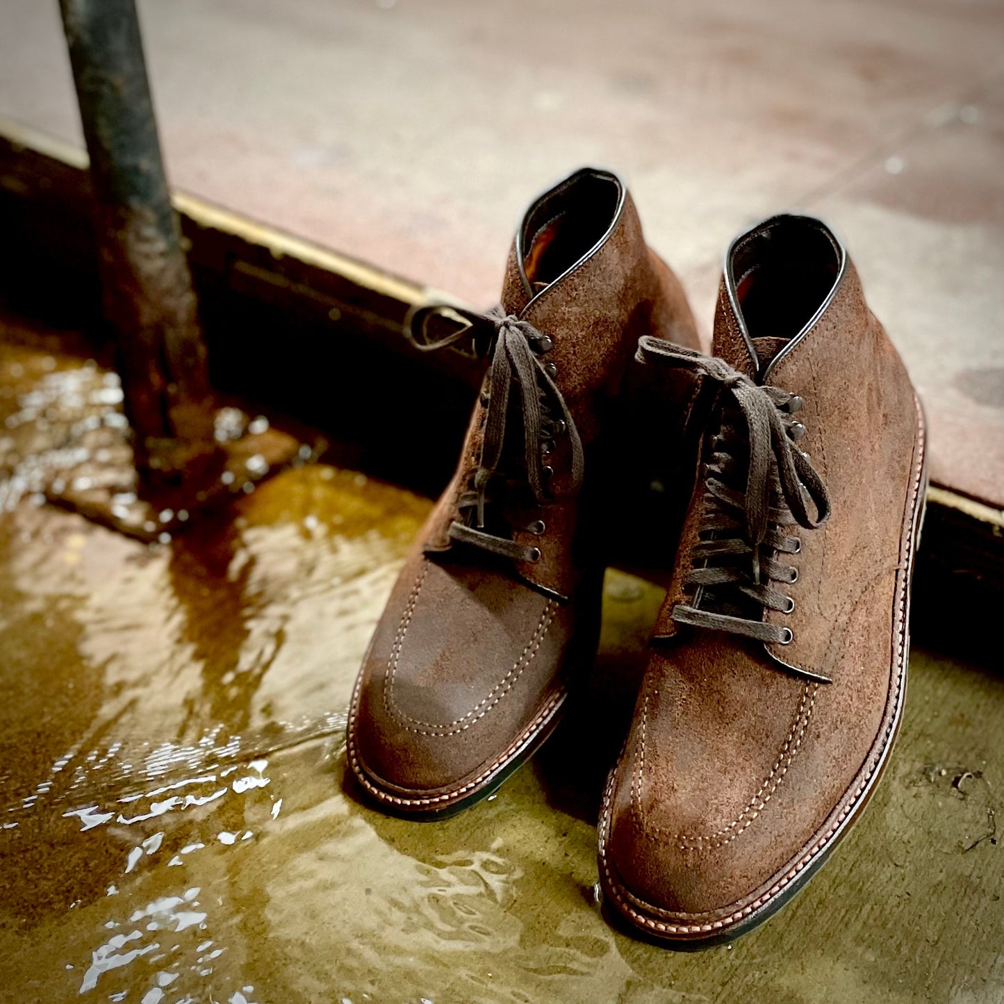 The Alden Madison 4015HC Indy Boot in Tobacco Reverse Chamois, featuring dark laces, sits on a wet, textured surface. Nearby, a puddle reflects the boots silhouette and a metal post against a slightly blurred background.