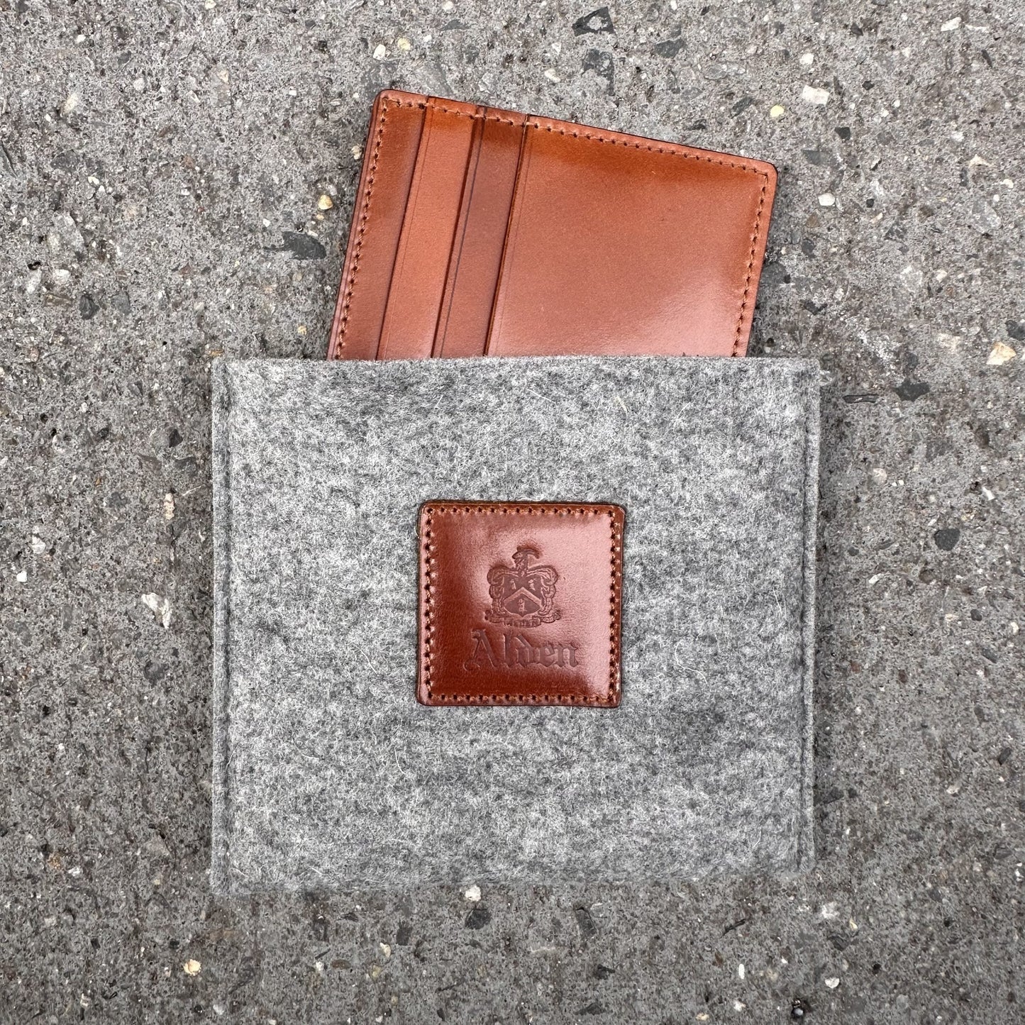 A brown leather LG 6005 - Alden Cognac Credit Card Holder with six pockets is partly nestled in a gray felt pouch with a leather patch labeled Allen, echoing the craftsmanship of Alden Madison, and rests elegantly on a textured gray surface.