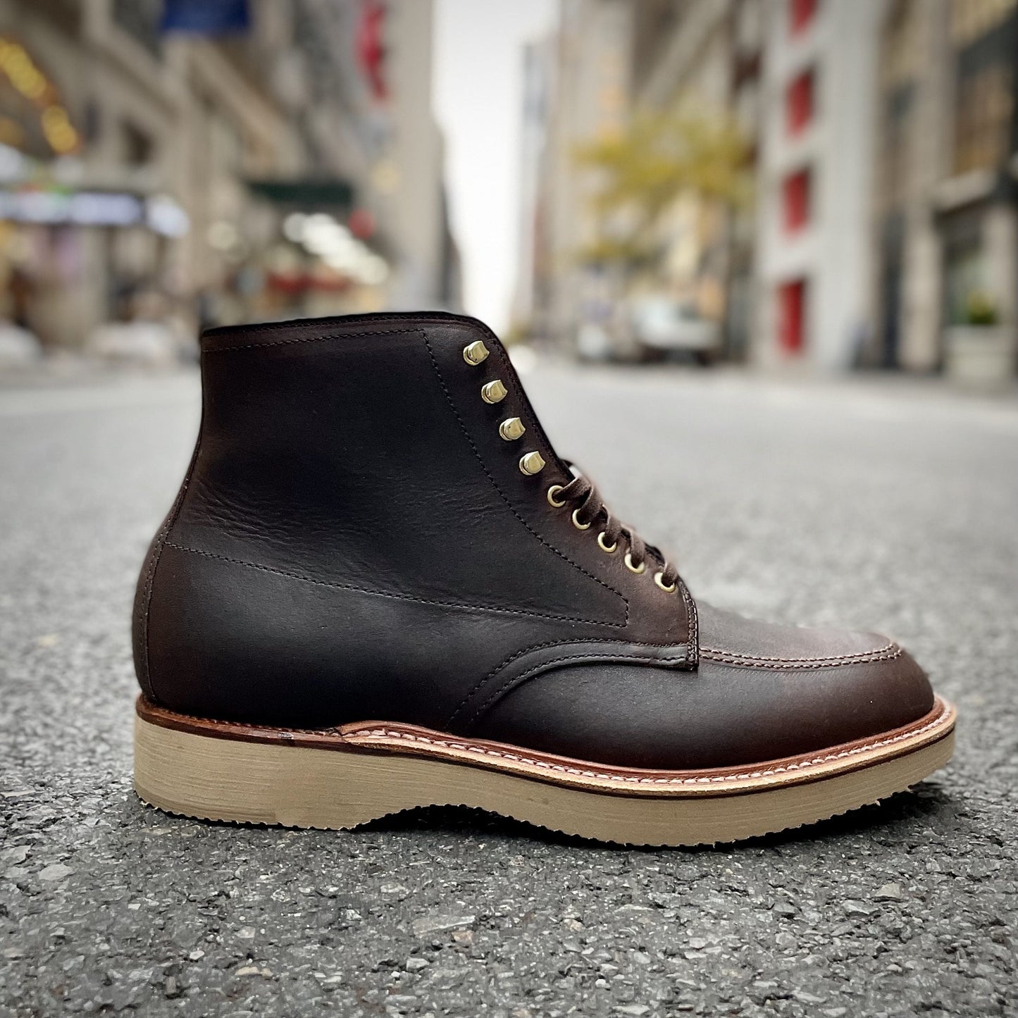 The Alden Madison D1926H - Porter Indy Boot, made from Kudu leather, features tan stitching, beige and textured soles on a city street. Gold eyelets contrast with blurred buildings and trees in the background.